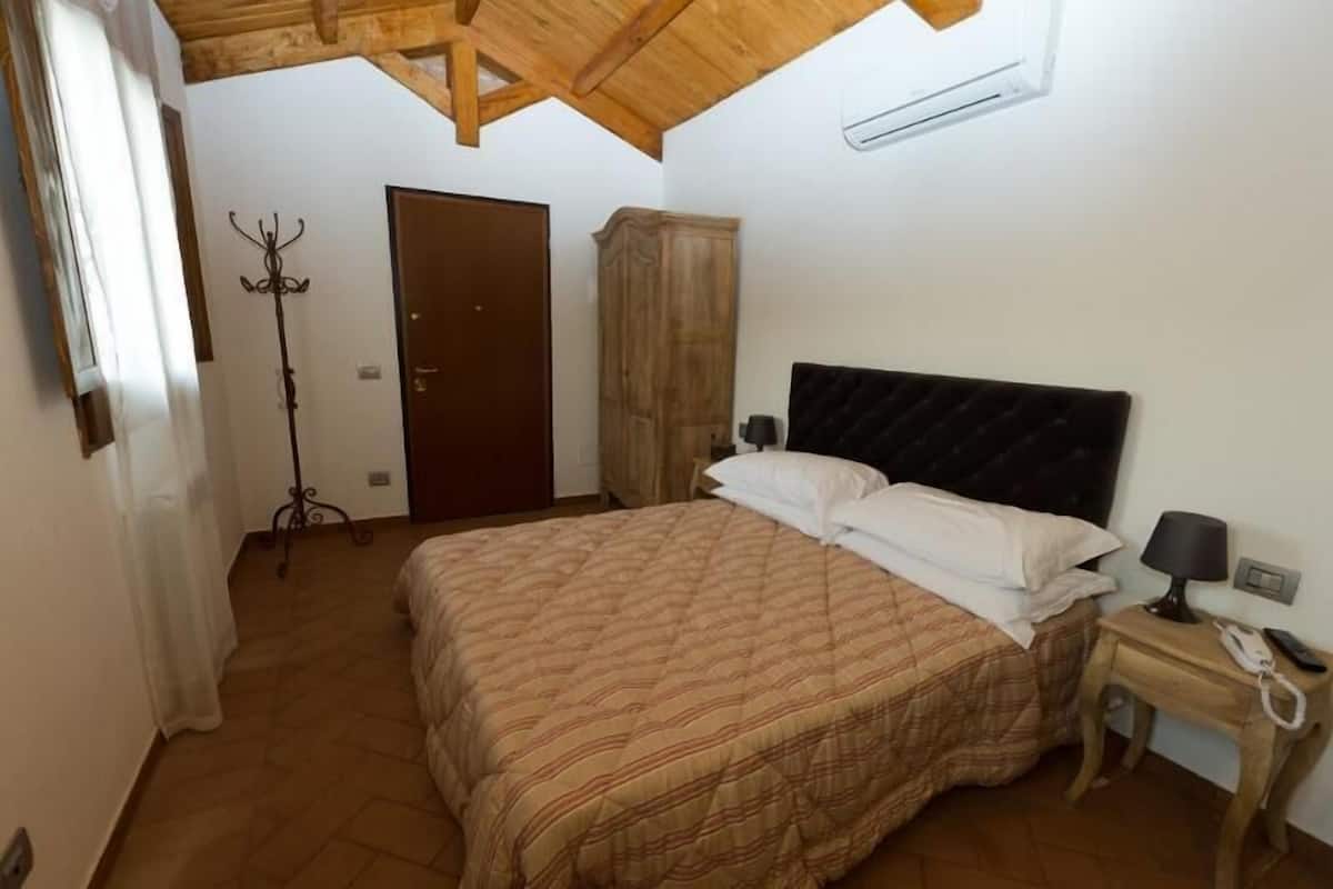 A bedroom with a bed, a wooden wardrobe, a coat rack, and a bedside table with a lamp and phone.
