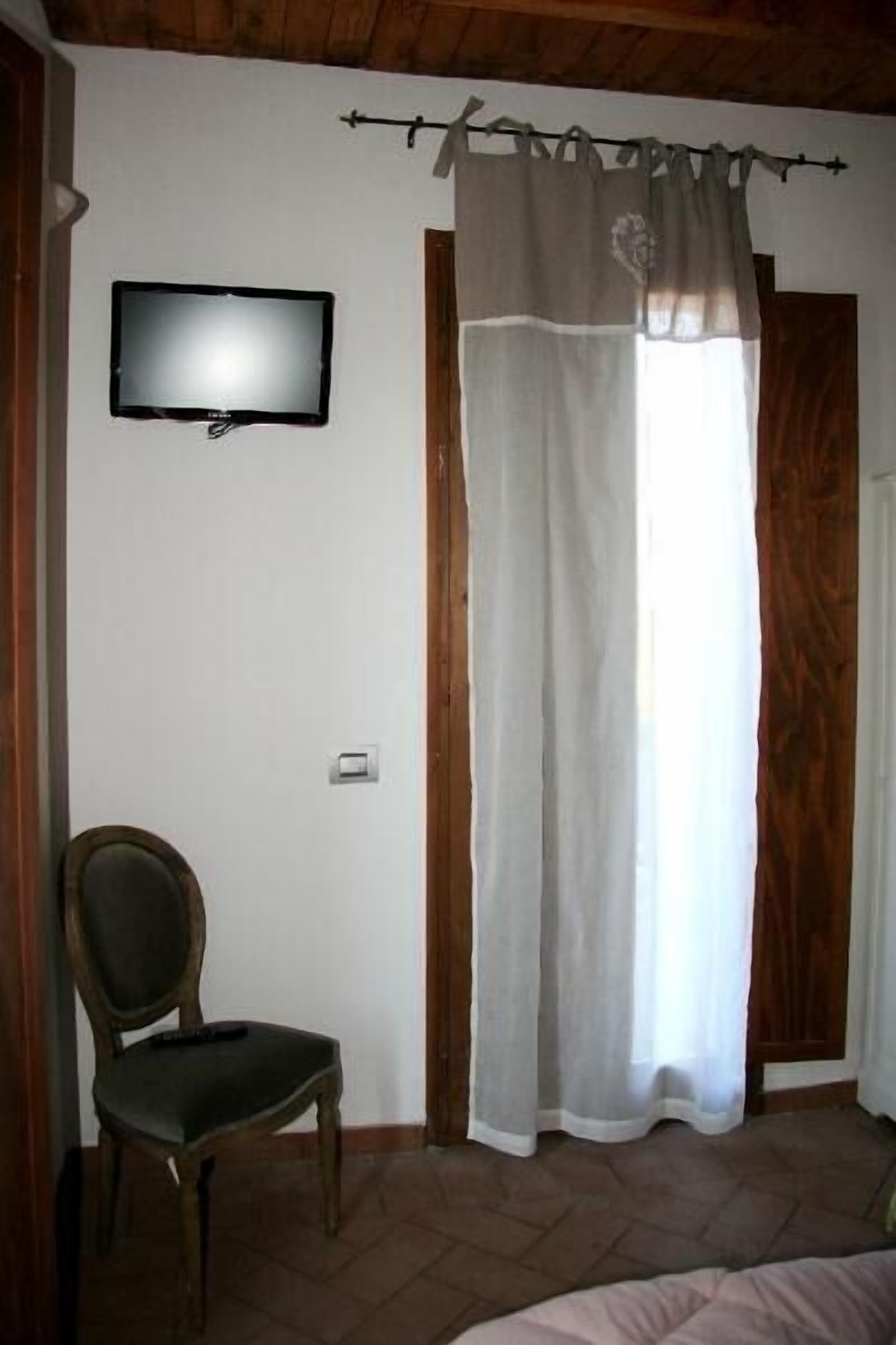 A room with a television mounted on the wall, a chair, a bed, and a window with a curtain.