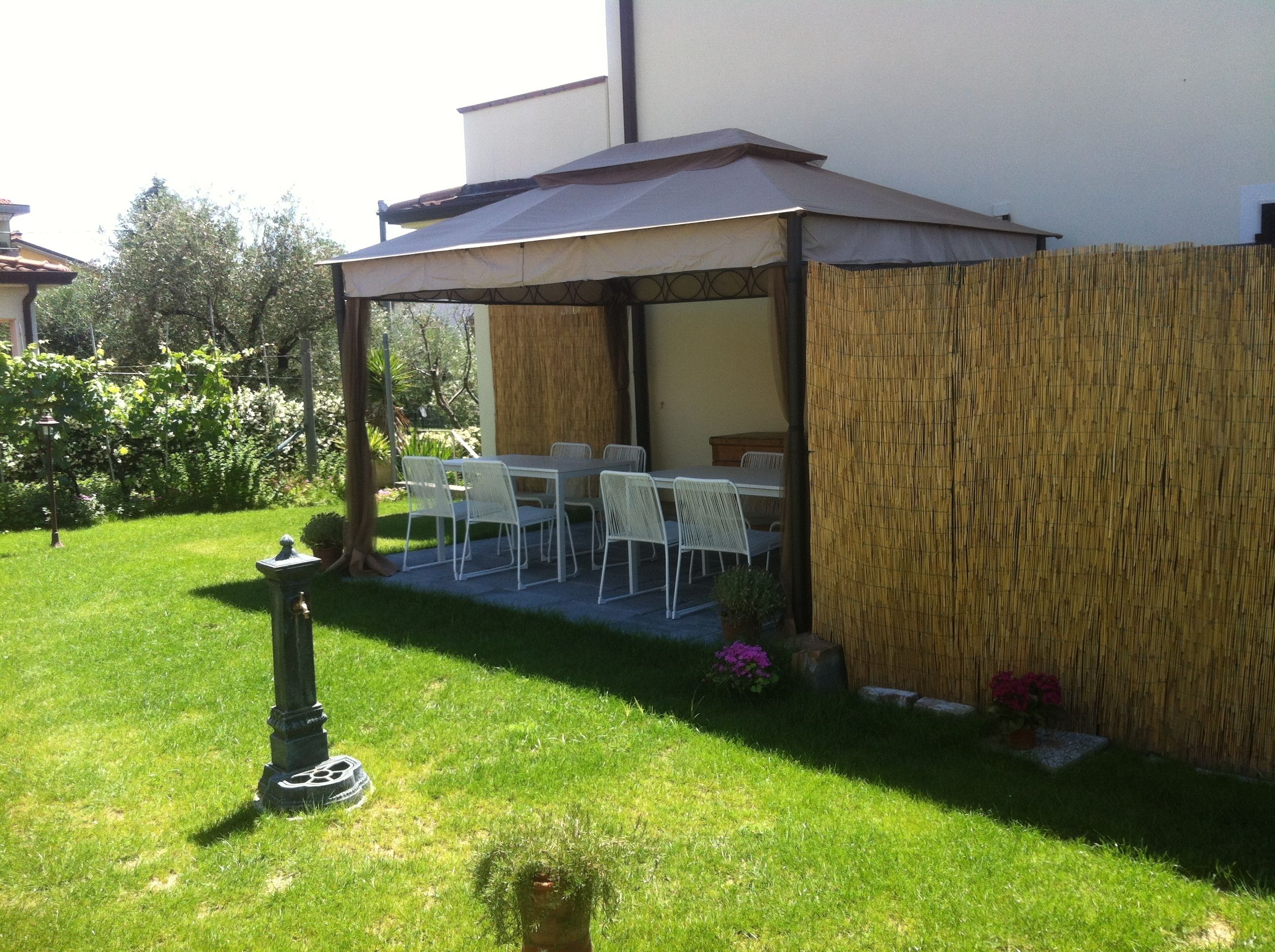 Un porche techado con una mesa y sillas, rodeado por una cerca de bambú, y un pequeño jardín con una fuente y plantas en macetas.