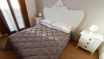 Ein ordentlich bezogenes Bett mit einer gesteppten, grauen Decke und weißen Kissen, ein weißer Nachttisch mit einer Lampe sowie eine weiße Kommode mit Schubladen.