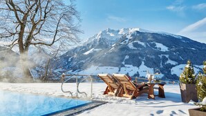 A snow-covered outdoor pool area with lounge chairs and a hot tub, set against a backdrop of snow-capped mountains.