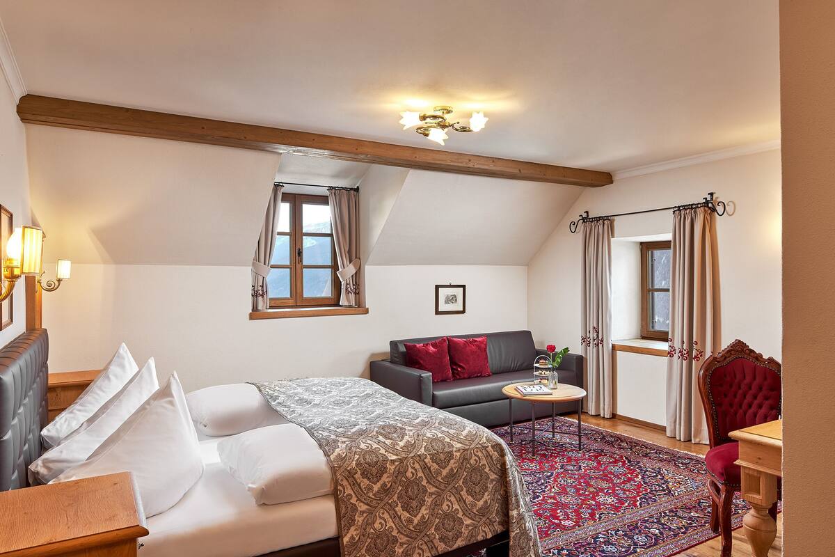 A bedroom with a bed, a sofa, a small table, and a chair. There is a window with curtains, a ceiling light, and a picture on the wall.