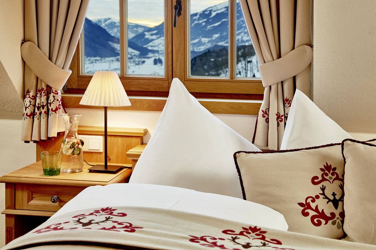 A hotel room with a bed, a nightstand, a lamp, and a view of mountains through the window.