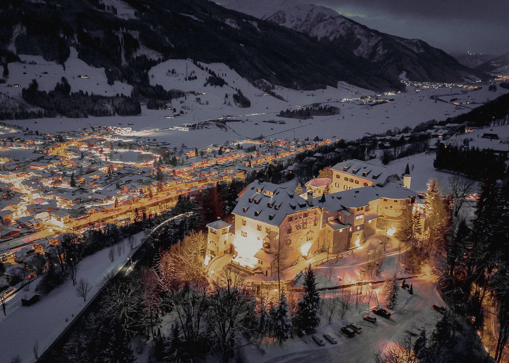 A snow-covered town with a prominent castle-like building in the foreground, surrounded by snow-covered hills and a lit-up town in the background.
