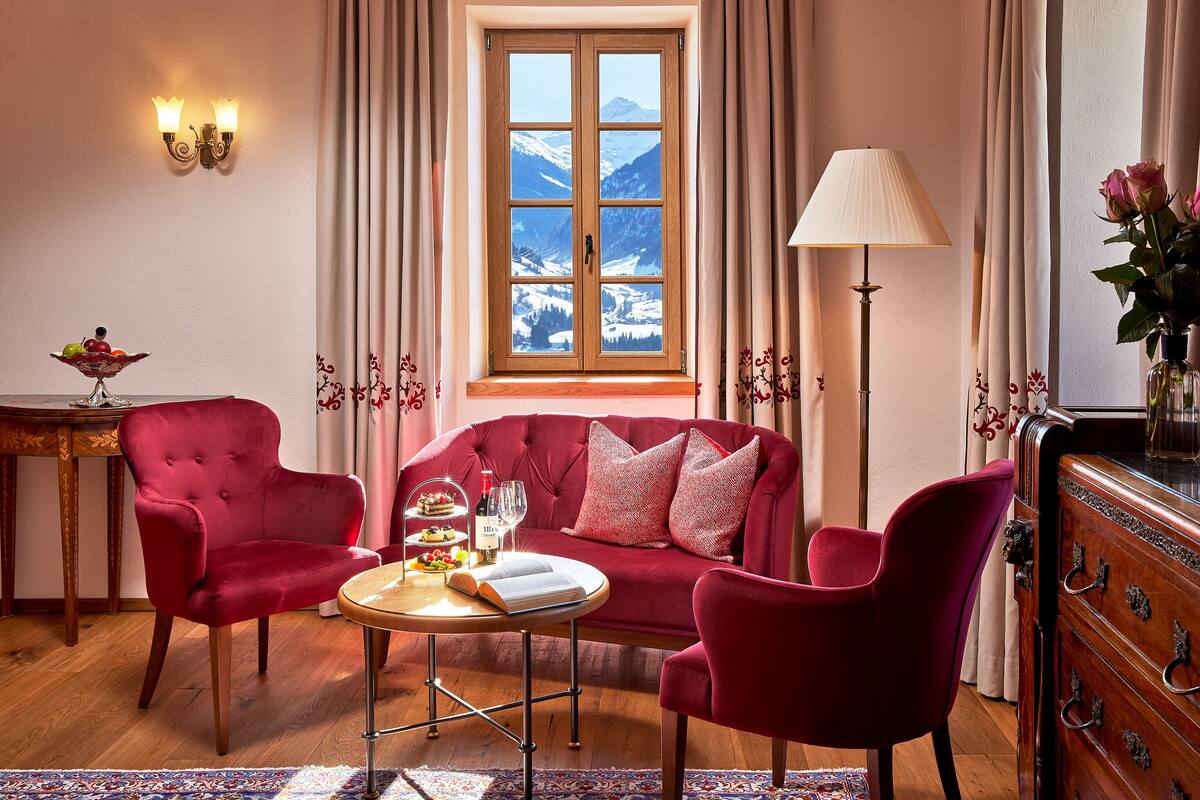 A cozy room with a red sofa, two matching armchairs, a wooden coffee table, and a view of snow-capped mountains through a large window.