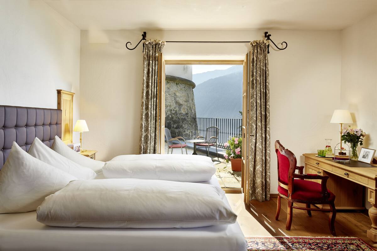 A hotel room with a bed, a desk, a chair, and a view of a mountainous landscape.