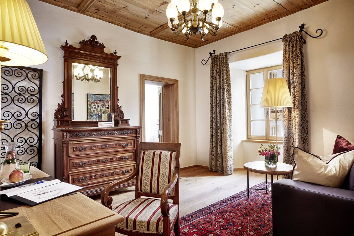 A room with a wooden dresser, a chair, a sofa, a chandelier, and a patterned rug.