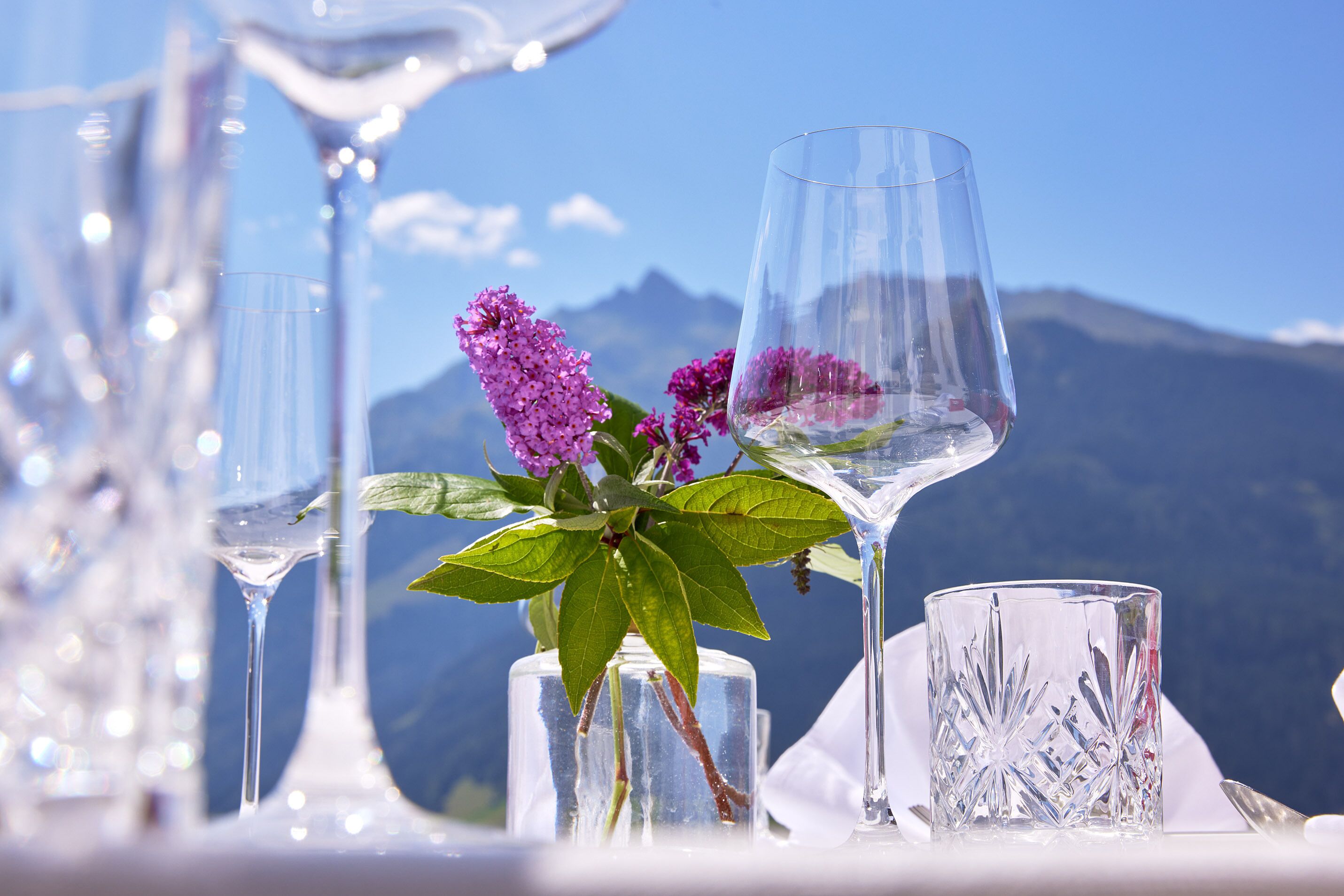 A table set with clear glassware and a vase with purple flowers against a mountainous backdrop.