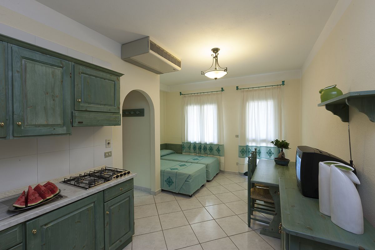 A compact kitchen with a gas stove, a dining table, and a small TV. There are two beds with matching green bedding in the adjacent room.