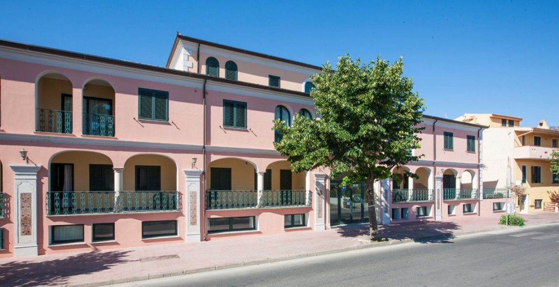 Un bâtiment rose doté de fenêtres en arc de cercle et de balcons, un arbre devant, et un ciel bleu clair.