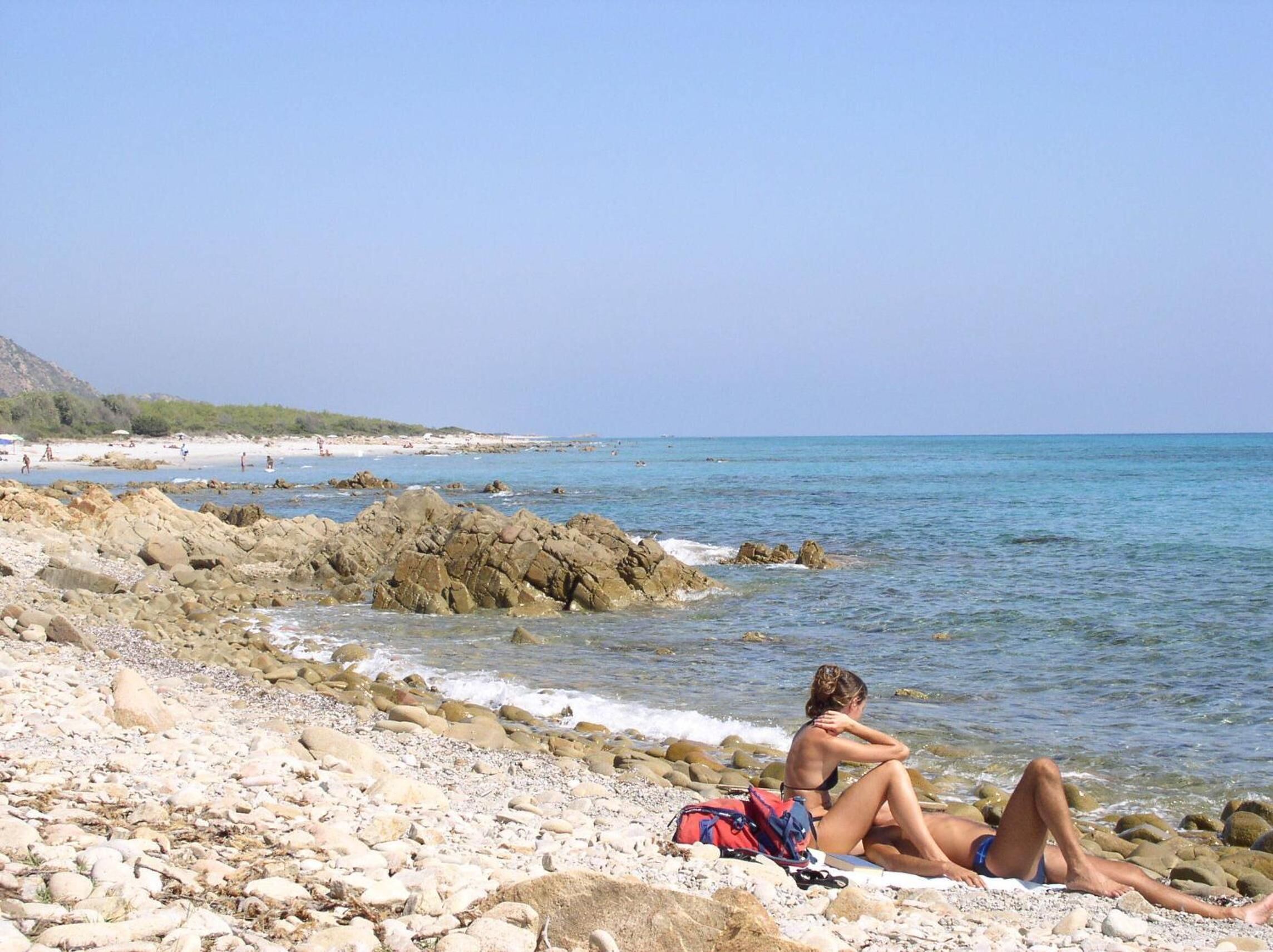 Una playa rocosa con una persona sentada sobre una toalla, una mochila y una vista despejada del mar y la costa lejana.
