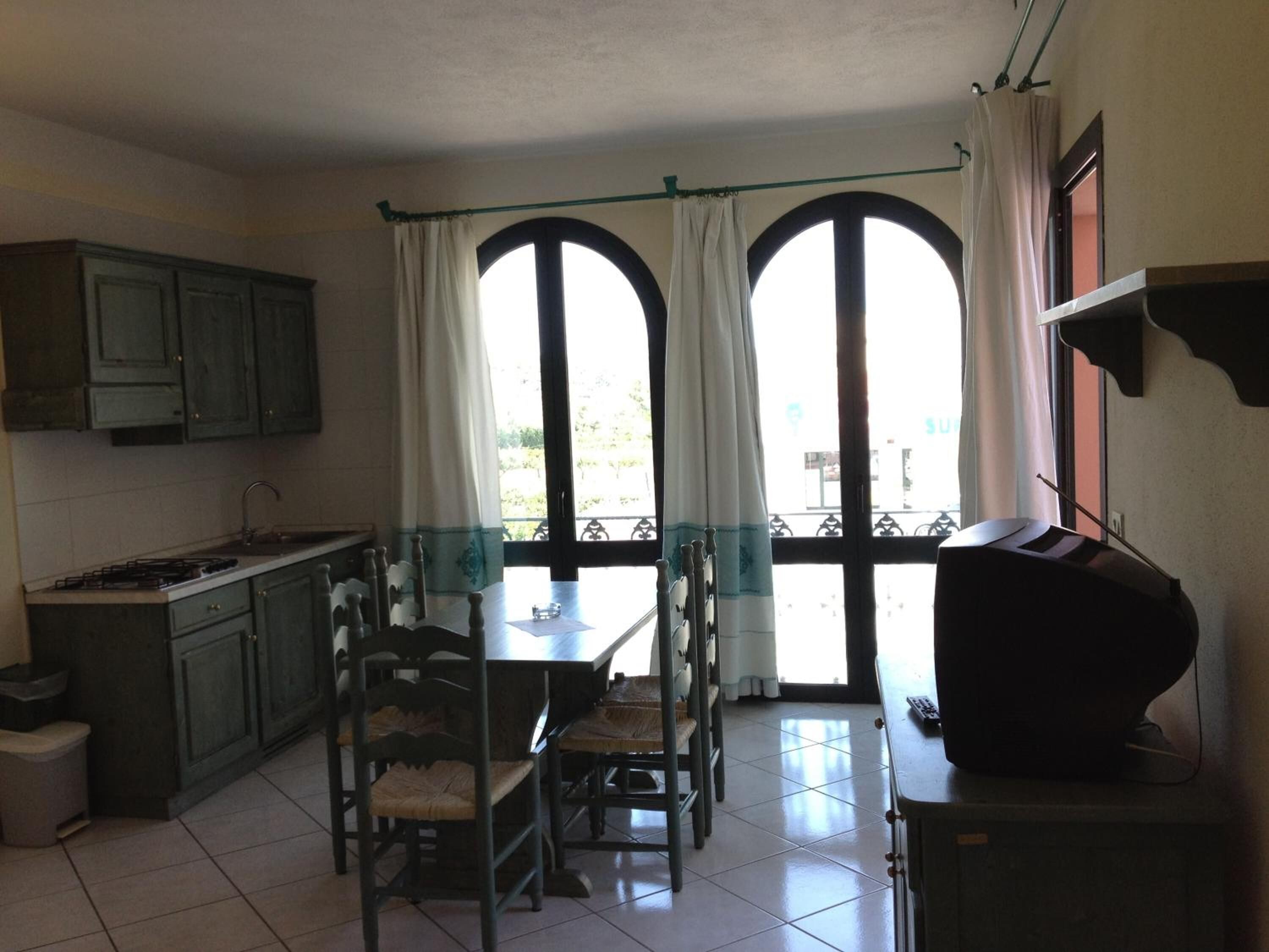 A kitchen with a dining area, featuring a table and chairs, a TV, and a view through large windows.