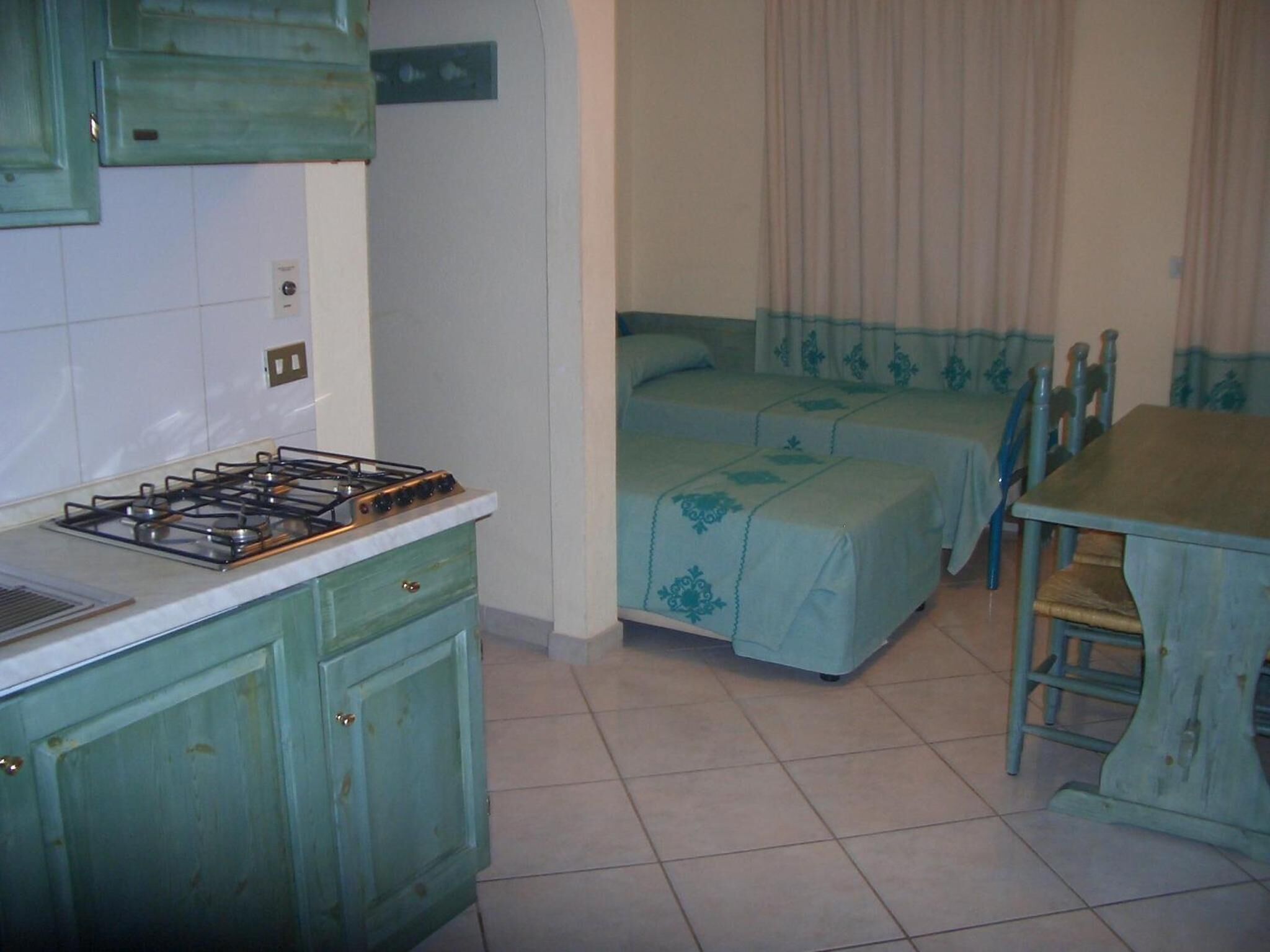 A small, compact kitchen with a gas stove, a single drawer, and a cabinet. There is a dining table with two chairs and a bed with a blue bedspread in the background.