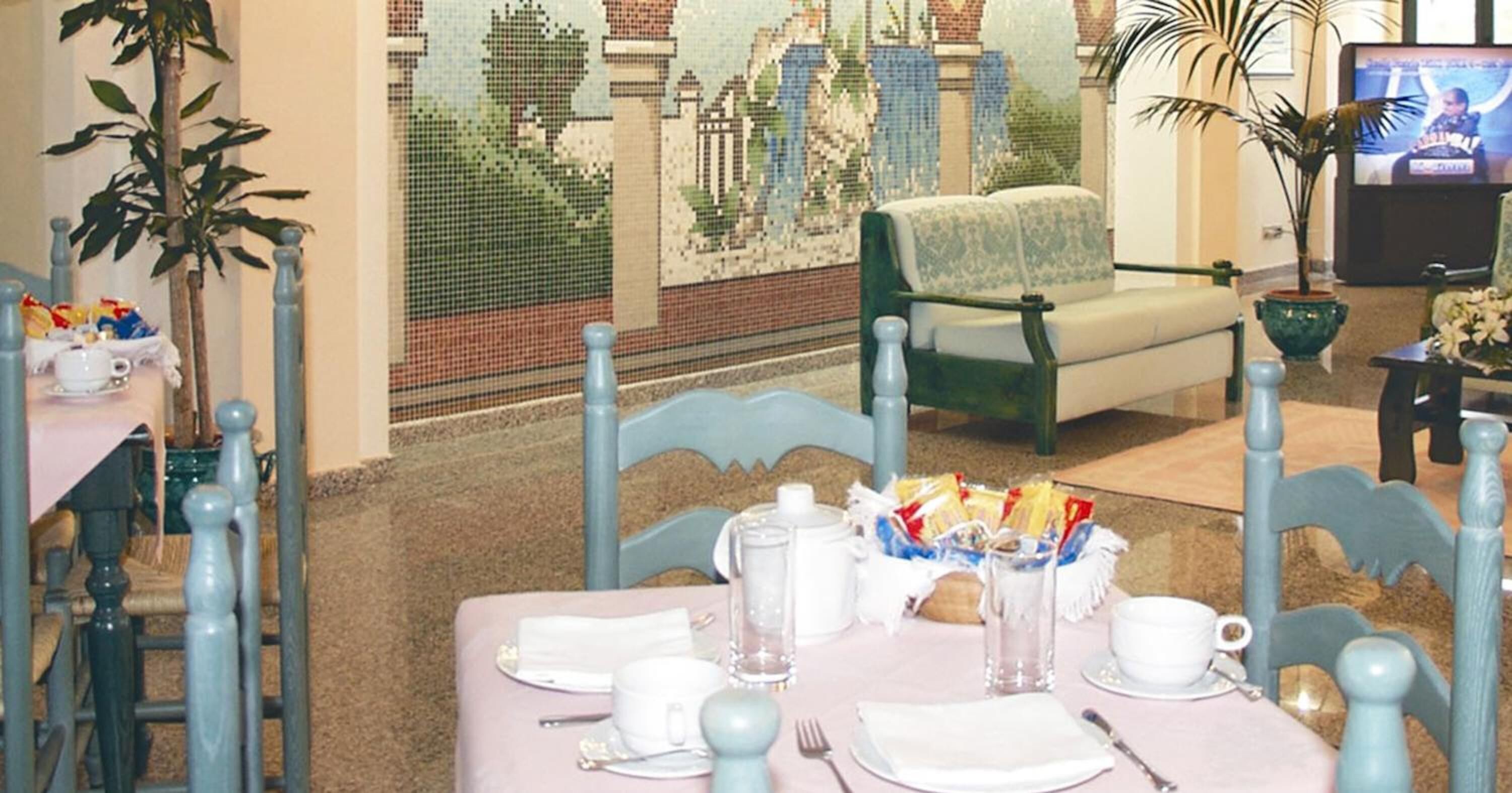 A dining area with a table set for two, chairs, a bench, and a television in the background.