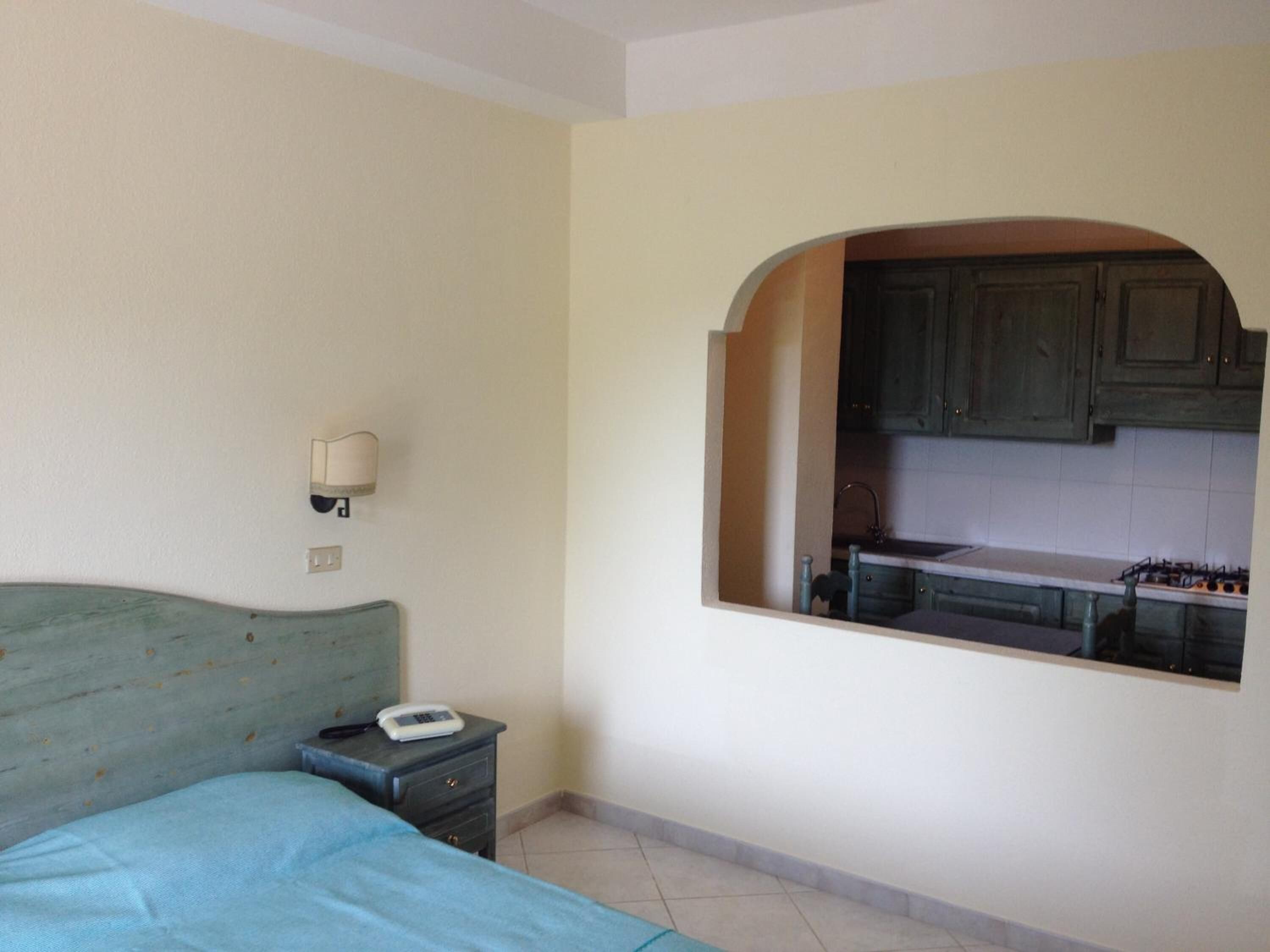Une chambre à coucher avec un lit, une table de chevet sur laquelle se trouve un téléphone, et une vue sur une cuisine par le biais d’une porte en arc de cercle.