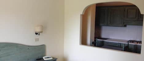 A bedroom with a bed, a bedside table with a phone, and a view of a kitchen through an arched doorway.