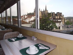 Un coin repas avec une table dressée pour deux personnes, une fenêtre donnant sur un bâtiment historique et un fauteuil en osier.