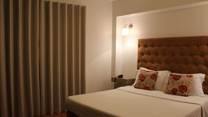 Une chambre d’hôtel avec un grand lit, une tête de lit, une table de chevet avec une lampe et une fenêtre avec des rideaux.