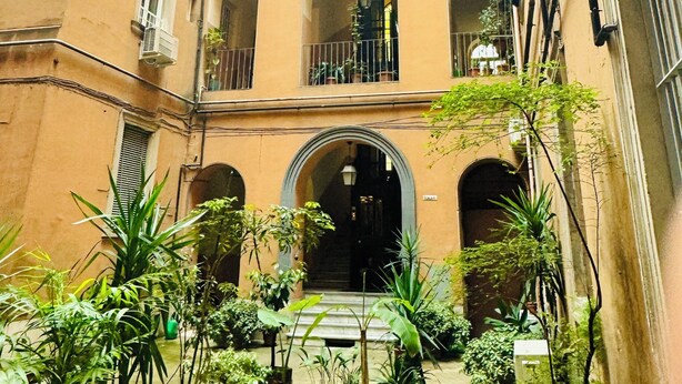 A multi-story building with balconies, a central courtyard with potted plants, and a staircase leading to the entrance. - Interno 5 (Rome)