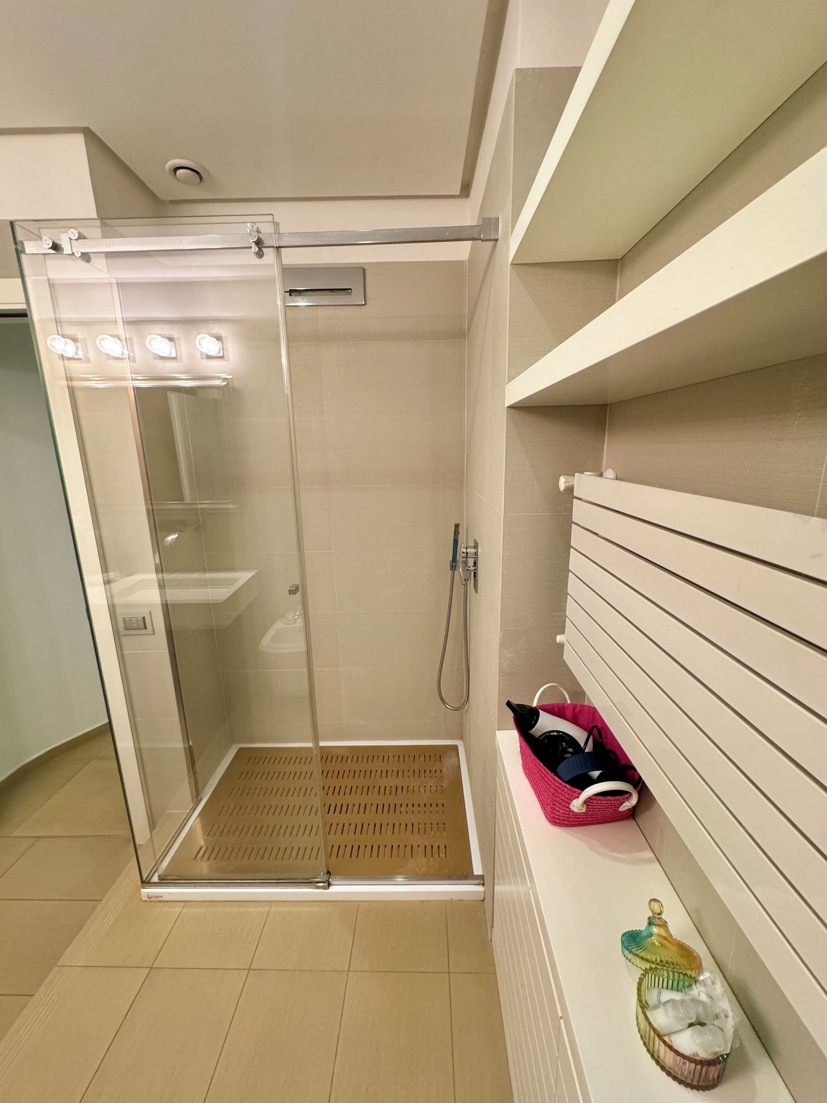 A glass-enclosed shower with a control panel, a white tiled floor, and a pink basket with items on a white shelf.