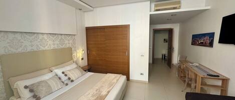 A bedroom with a large bed, a wooden wardrobe, a desk with a chair, and a television on the wall.