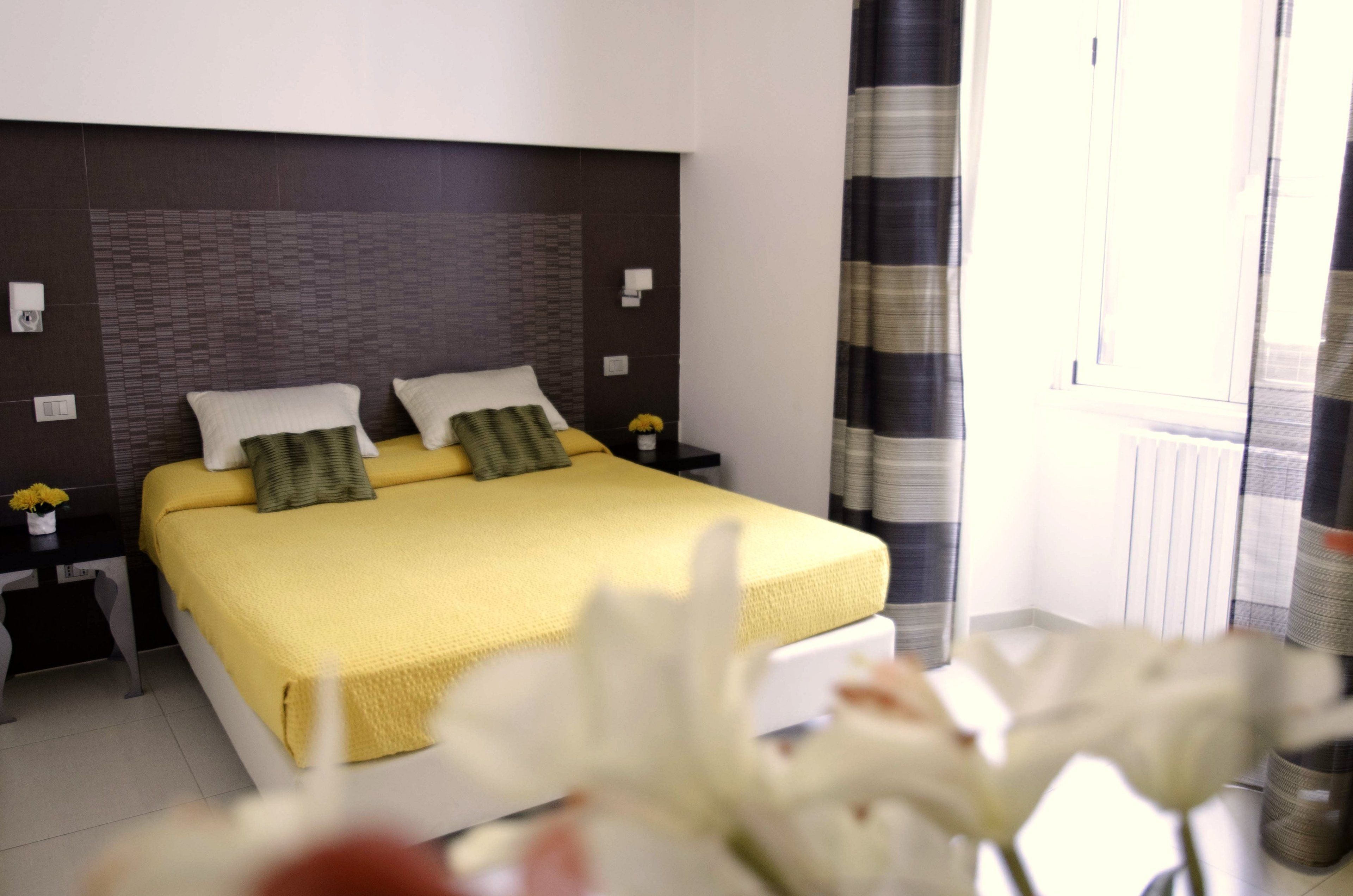 A hotel room with a bed, nightstands, a window with curtains, and a vase with flowers.