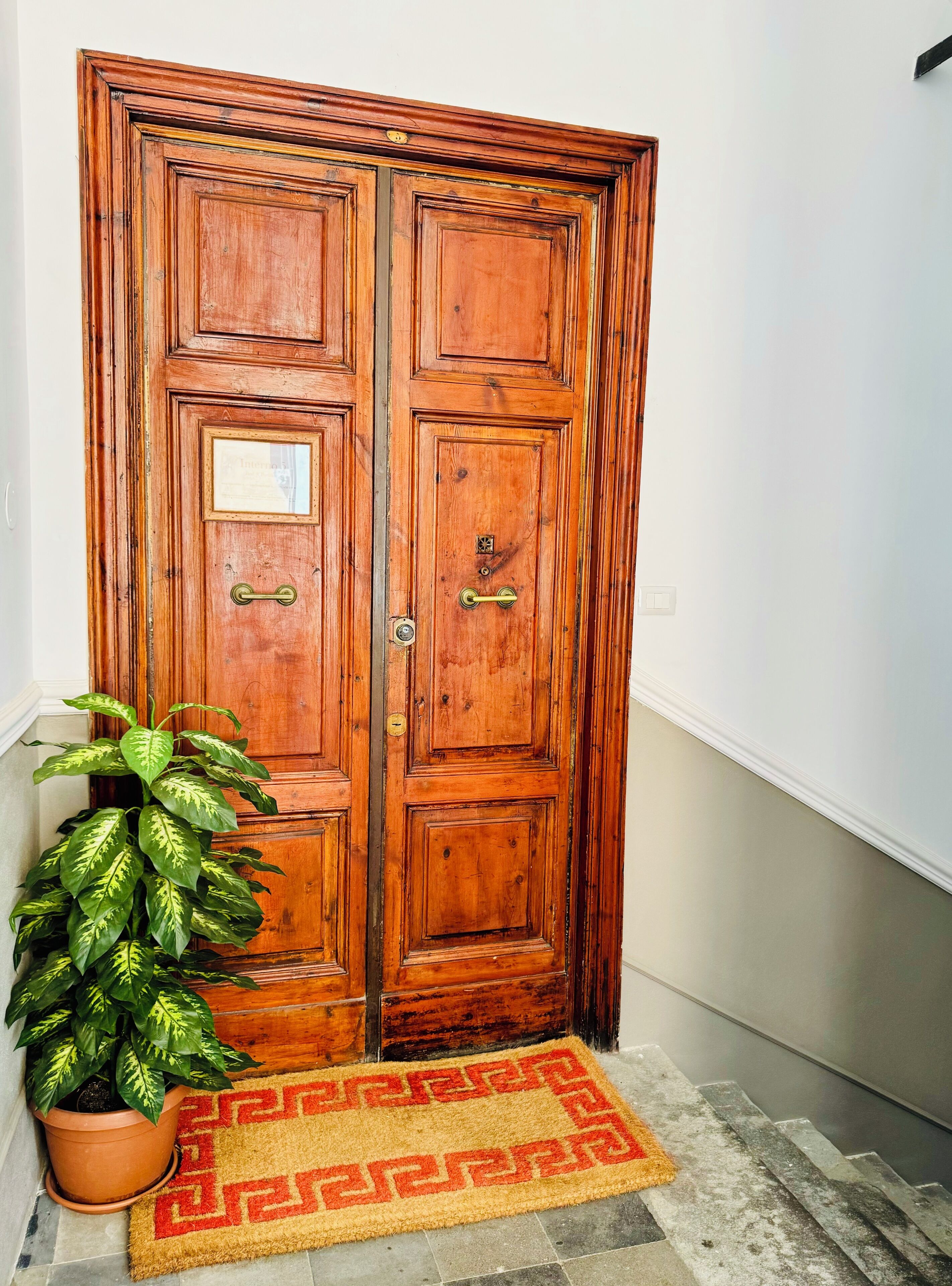 Une porte à deux battants en bois, dotée d’une poignée en laiton et d’une petite plaque, un pot de fleurs et un paillasson à motifs.