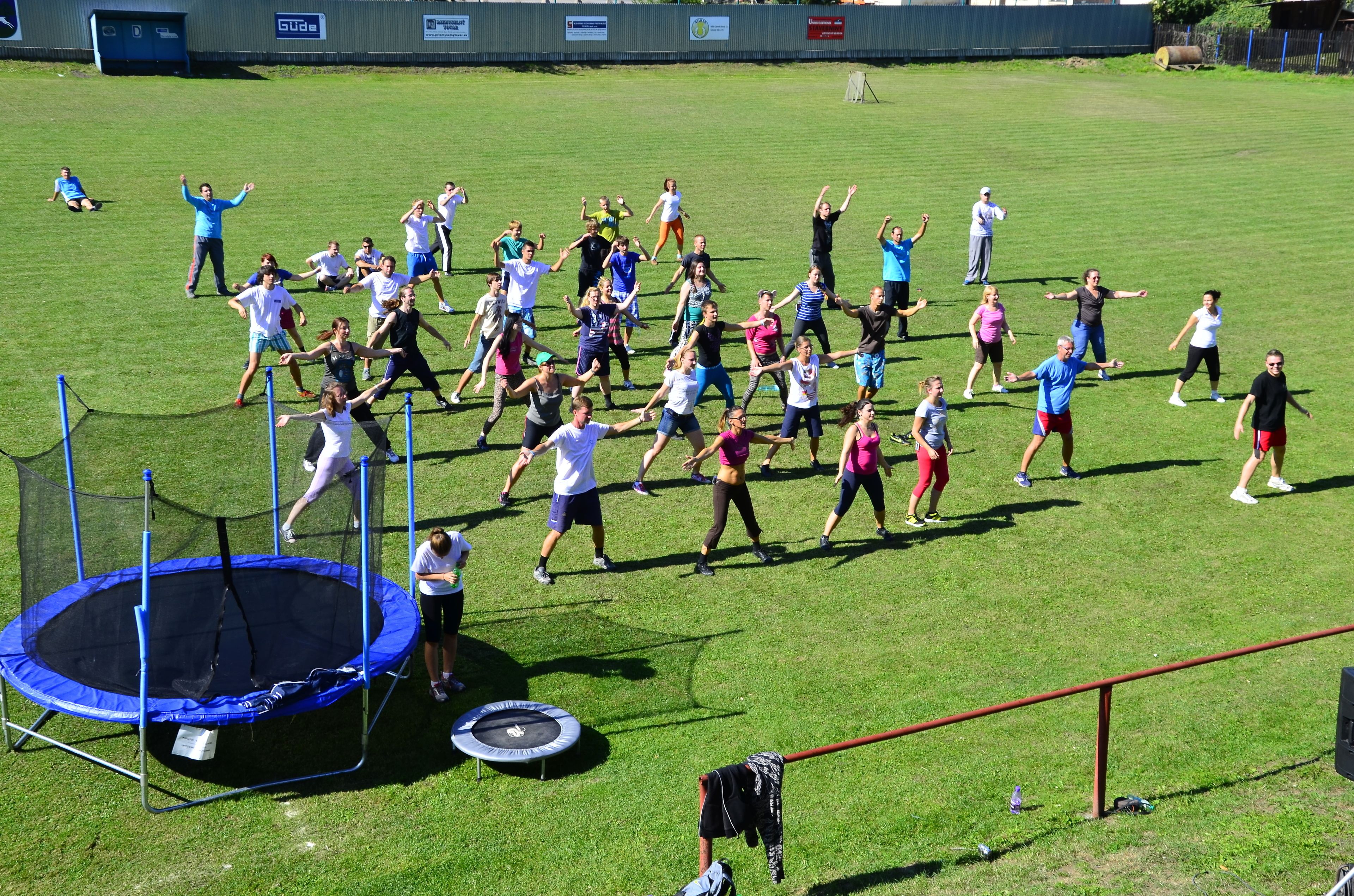 Un grupo de personas haciendo ejercicio al aire libre en un campo cubierto de césped, con un trampolín y una cerca roja al frente.