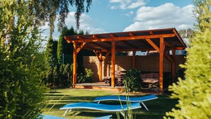 Een houten pergola met dak, een overdekte zithoek en twee blauwe lounge fauteuils op een gazon.