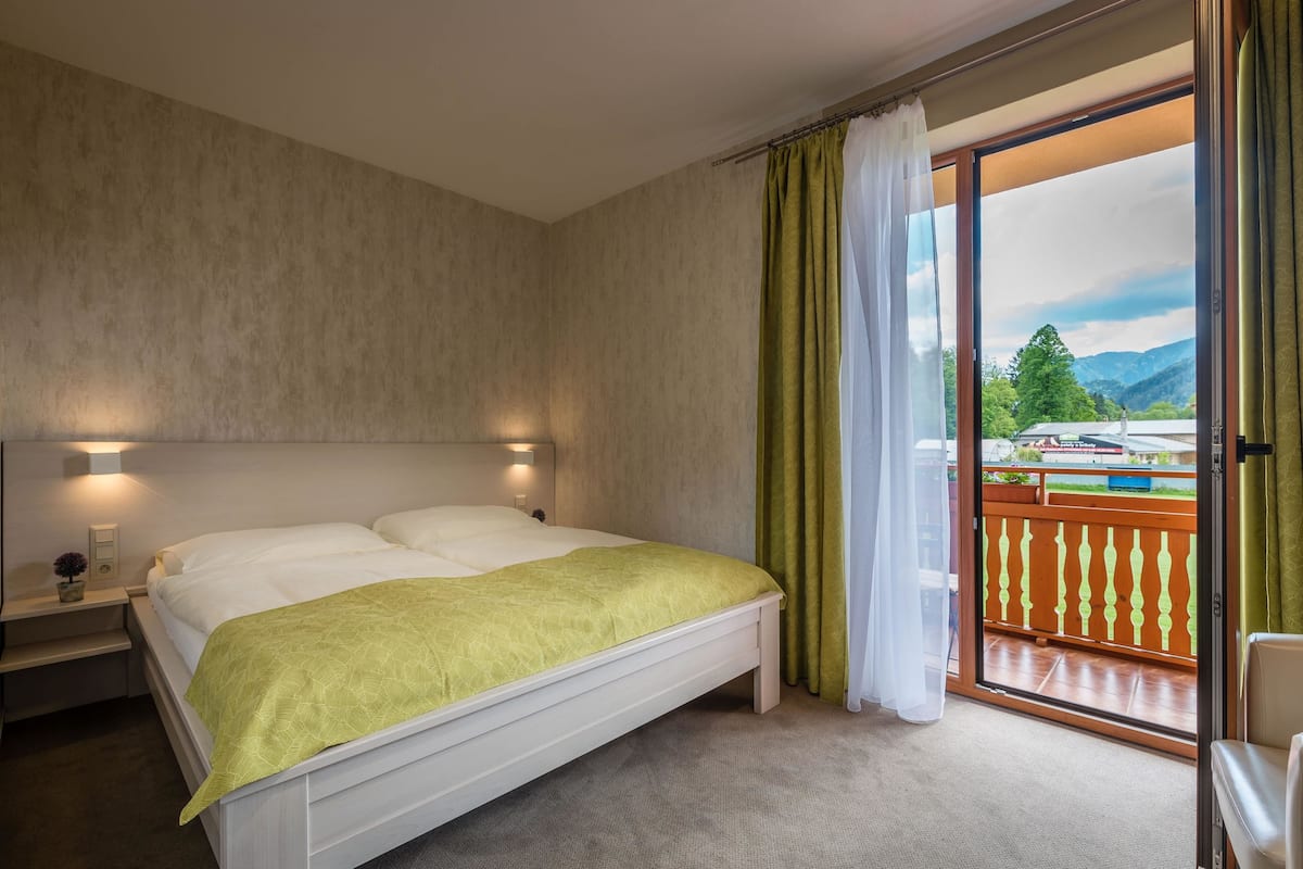 A bedroom with a bed, bedside tables, a nightstand, a green blanket, and a view of a balcony with a railing and greenery.