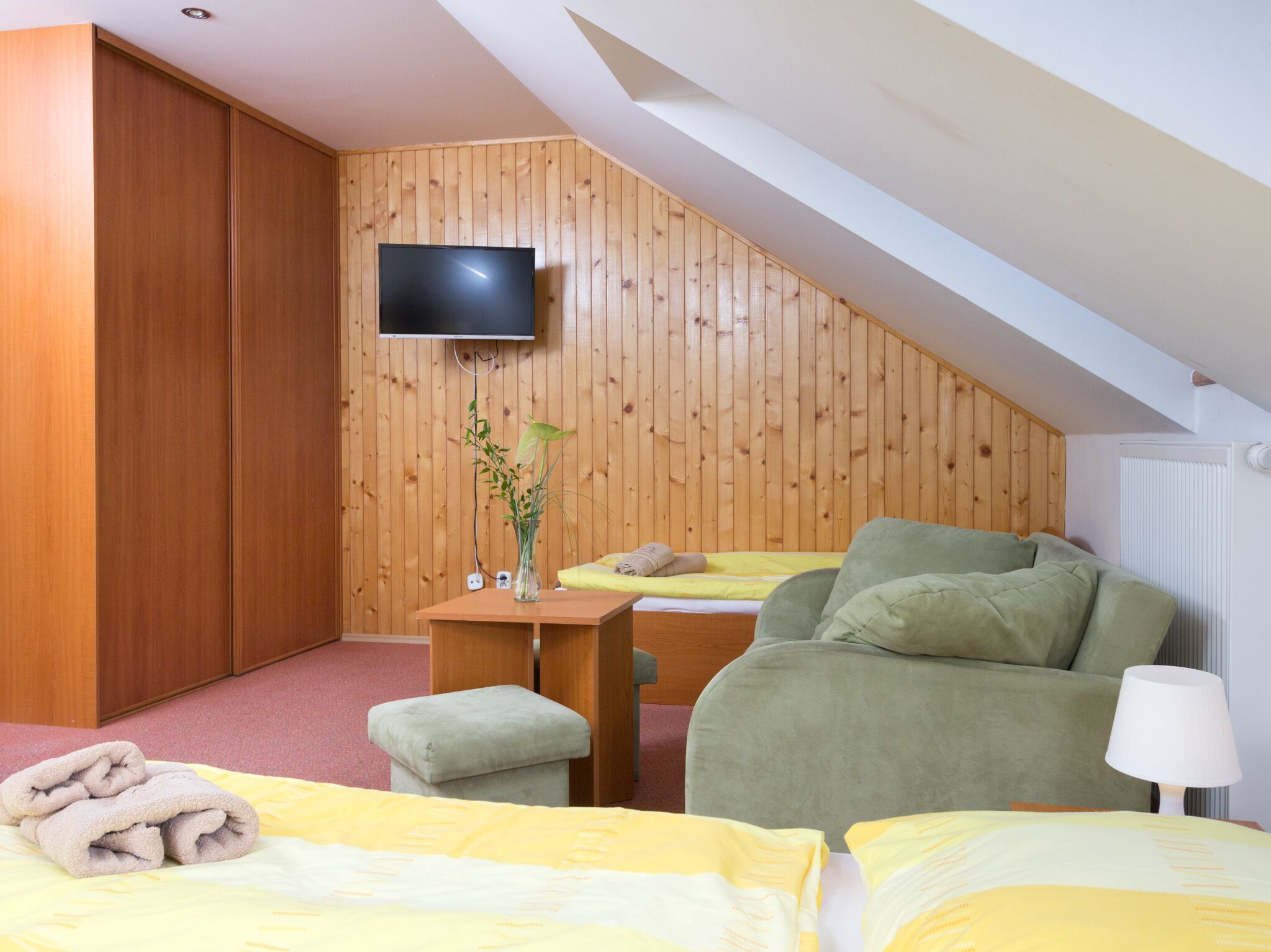 A bedroom with a sloped ceiling, wooden walls, a bed with yellow bedding, a green armchair, a small table with a vase, and a TV mounted on the wall.