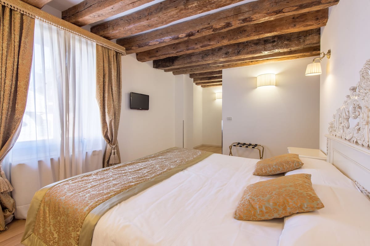 Ein Schlafzimmer mit Holzbalkendecke, einem Bett mit weißer und gemusterter Bettwäsche sowie einem Fernseher an der Wand.
