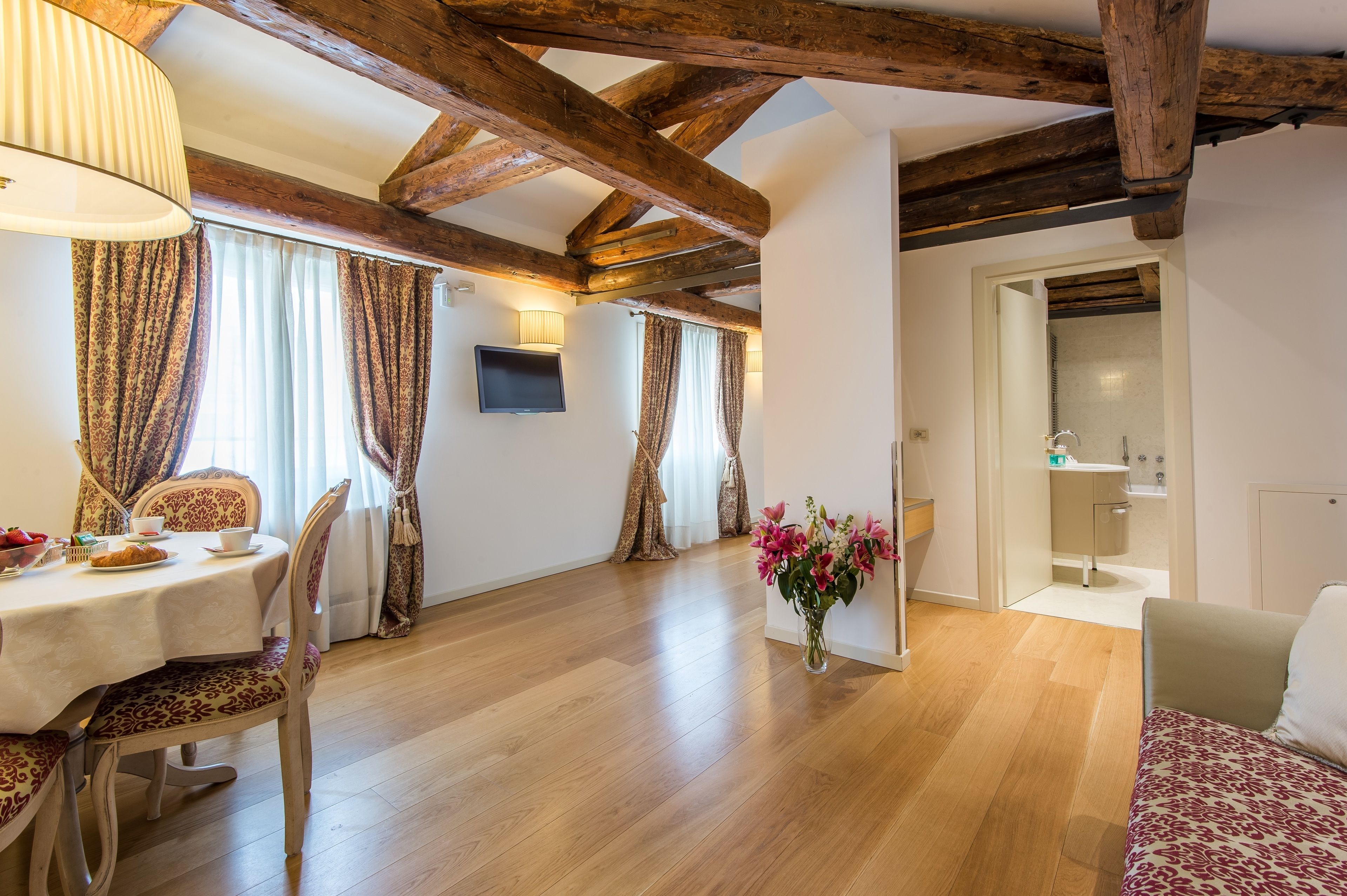 Un coin repas avec une table en bois, des chaises et un vase de fleurs. Un téléviseur est fixé au mur et on aperçoit une salle de bain à travers une porte ouverte.