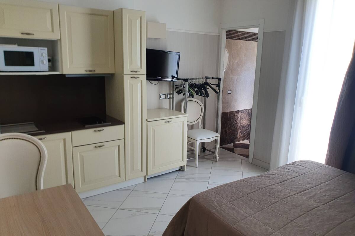 A compact hotel room with a kitchenette, a bed, a chair, and a TV.