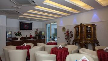 Un vestíbulo de hotel con varias mesas redondas cubiertas con manteles rojos, sillas color beige, un aparador de madera y un televisor mostrando el logotipo de RTL.