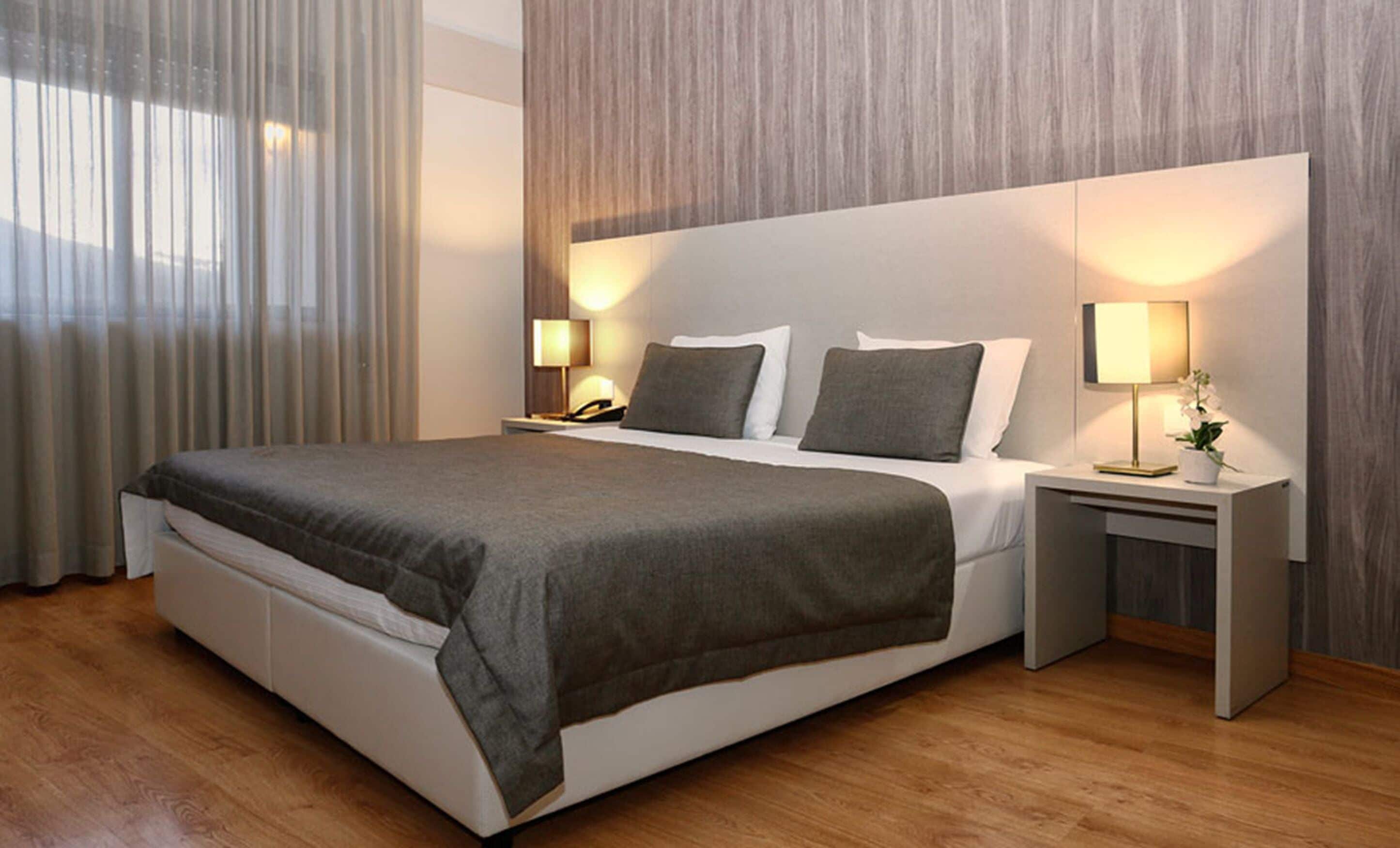 Habitació d'hotel moderna amb un llit gran, dues taules de nit amb làmpades i vista a l'exterior a través de cortines translúcides.