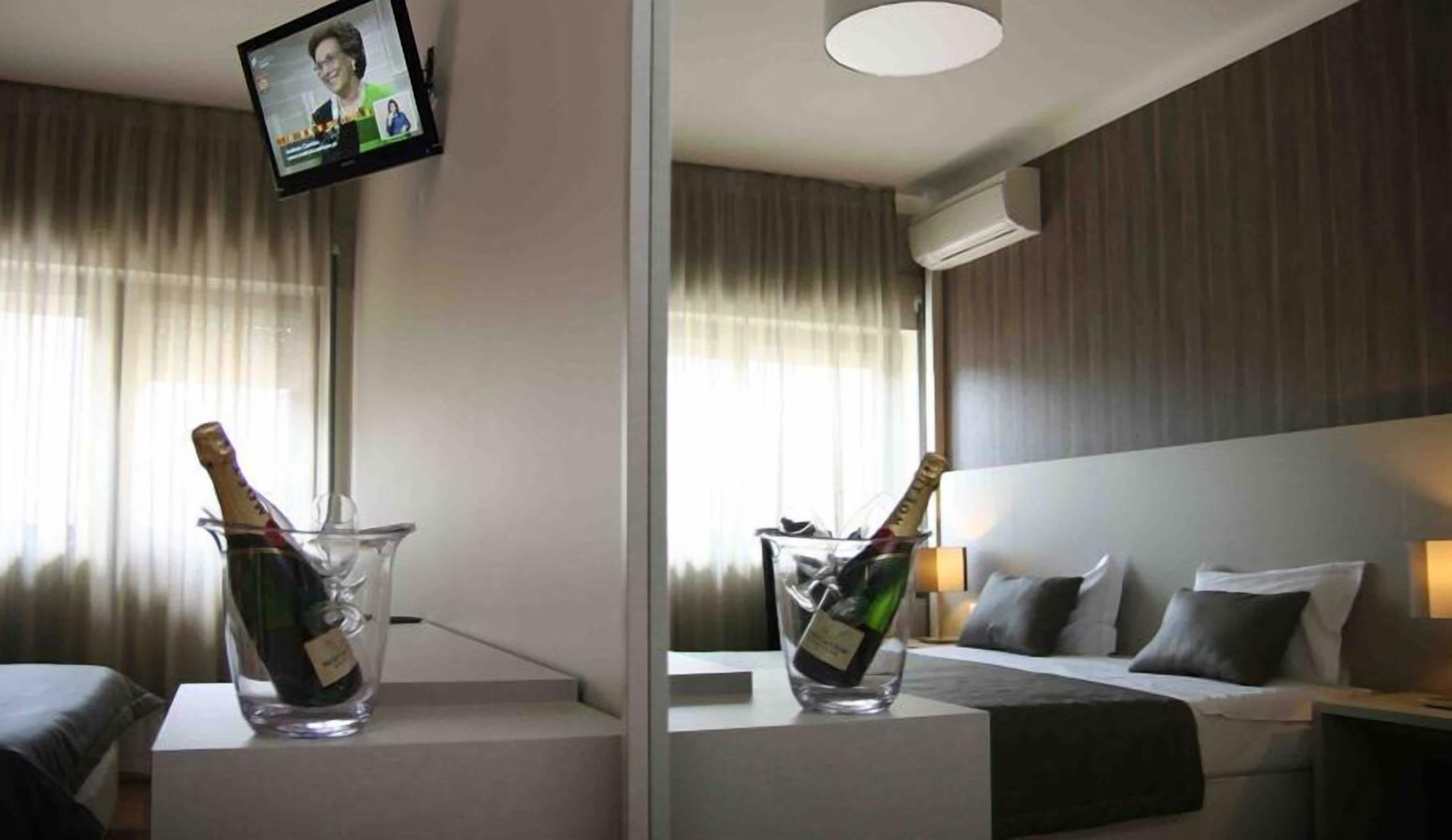 Una habitación de hotel con una cama, una mesita de noche con una botella de champán, un televisor montado en la pared y una ventana con cortinas.