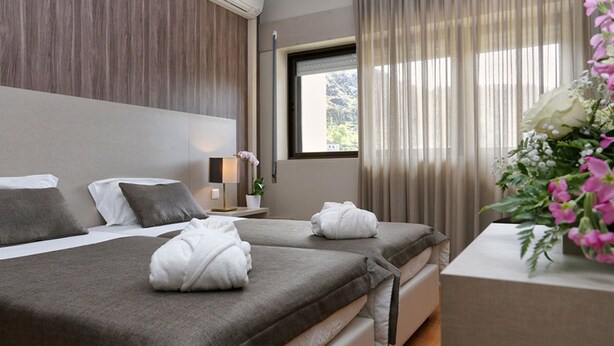 A hotel room with two beds, a window with curtains, a bedside table with a lamp, and a vase with flowers. - Hotel Douro Inn (Tabuaço)