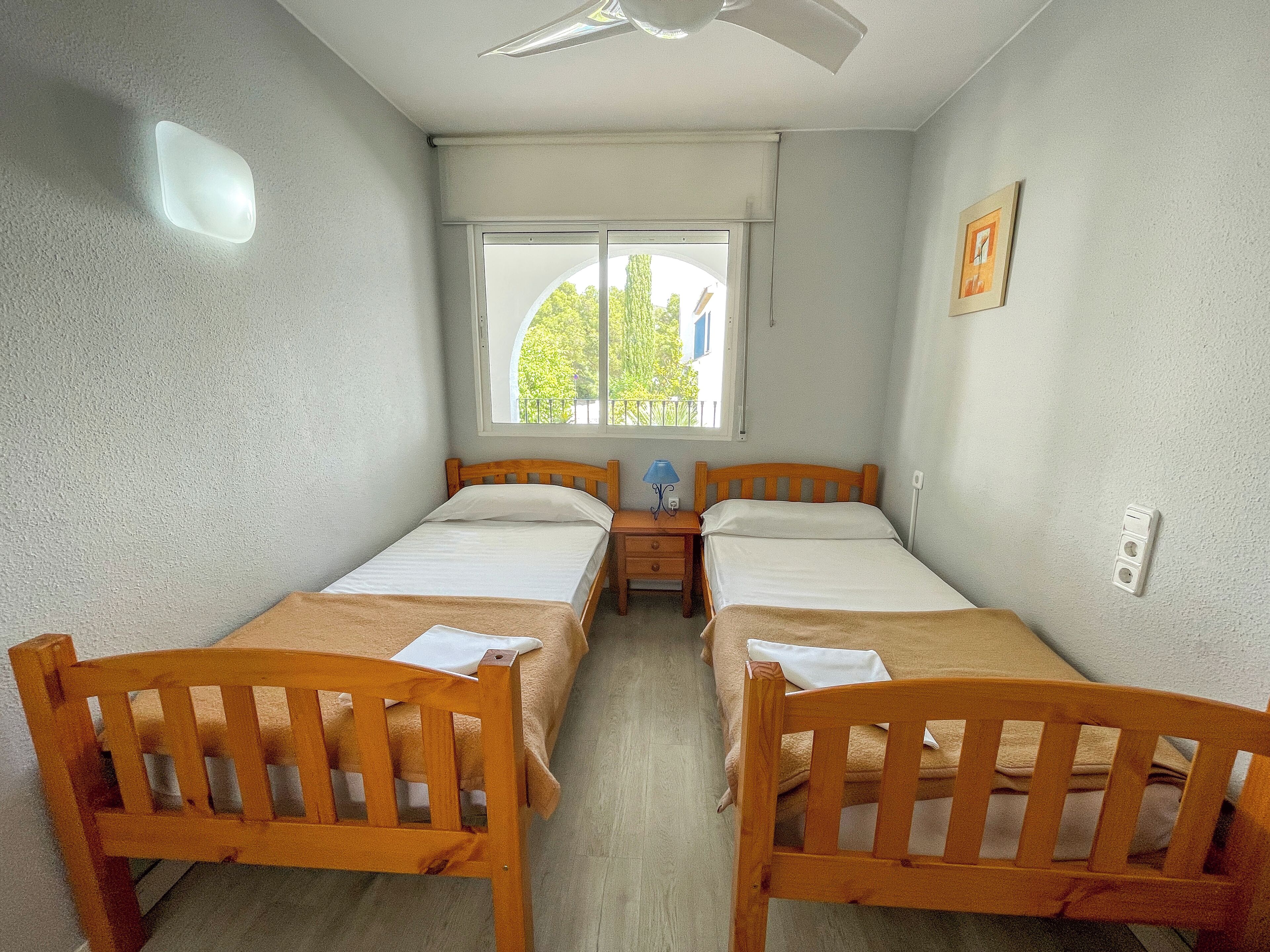 Una habitación pequeña y limpia con dos camas de madera, una ventana que da a árboles, un ventilador de techo y una mesita de noche con una lámpara.