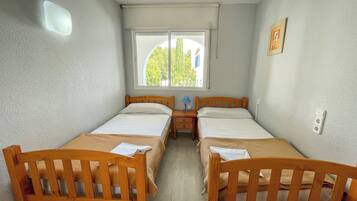 Una habitación pequeña y limpia con dos camas de madera, una ventana que da a árboles, un ventilador de techo y una mesita de noche con una lámpara.