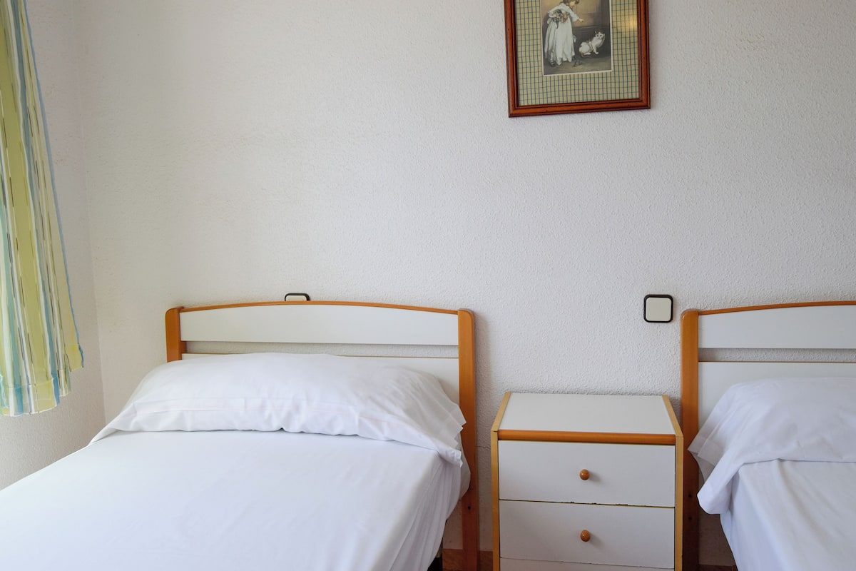 Habitación doble con dos camas individuales, una mesita de noche pequeña con cajones y un cuadro enmarcado en la pared.