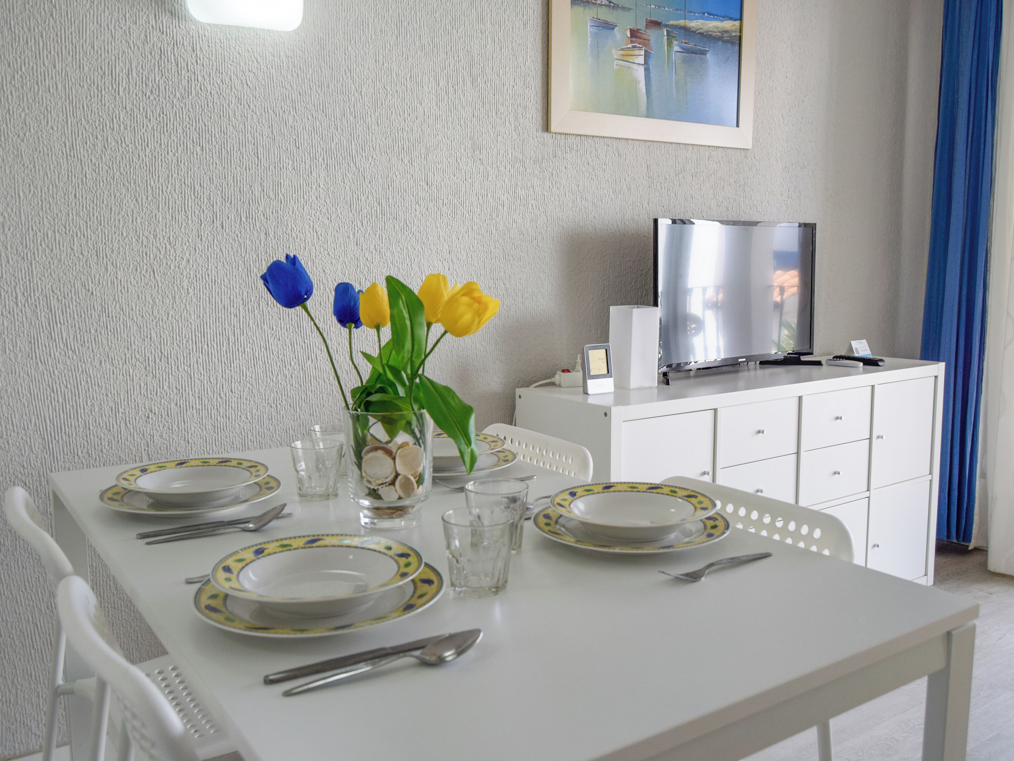 A dining table set with plates, cutlery, and a vase with flowers. A television and a small table are visible in the background.