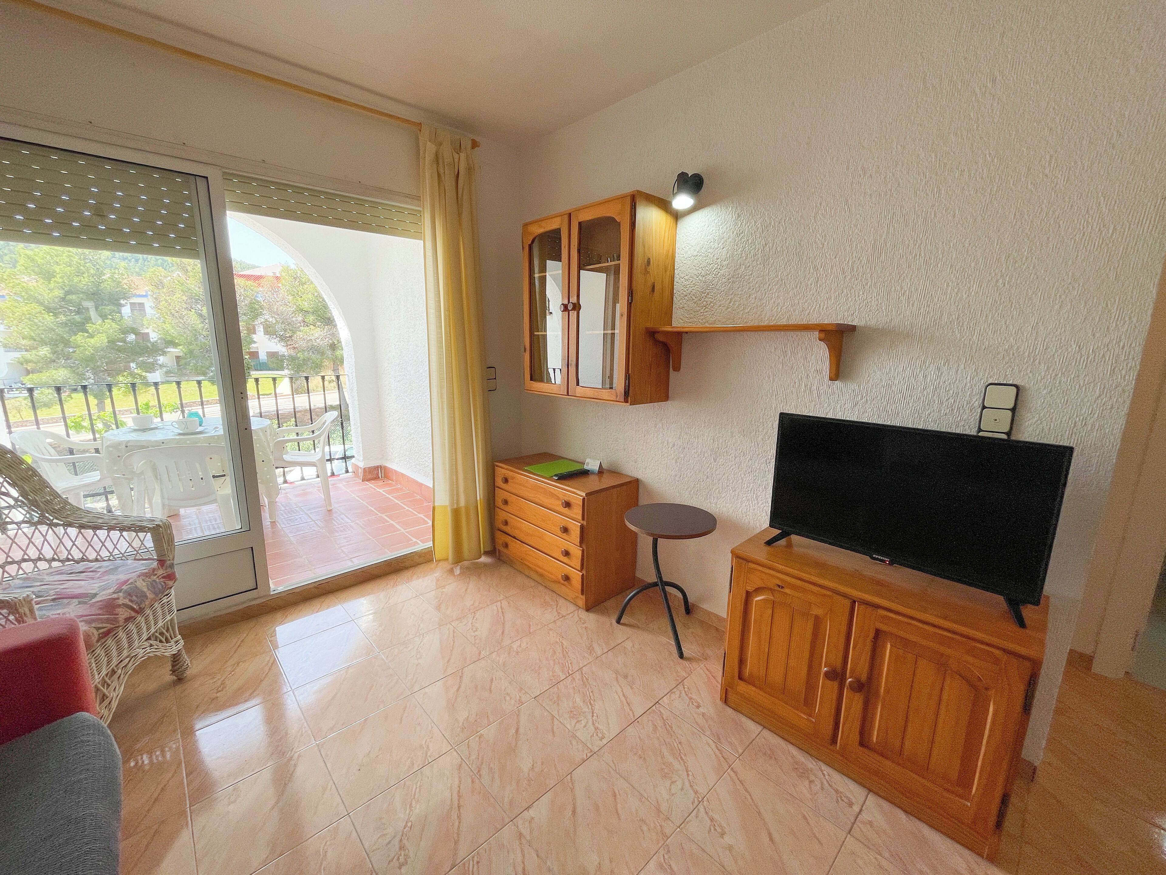 Una habitación con puerta de cristal corredera que da a un balcón, un mueble de madera con televisor y una mesita redonda.