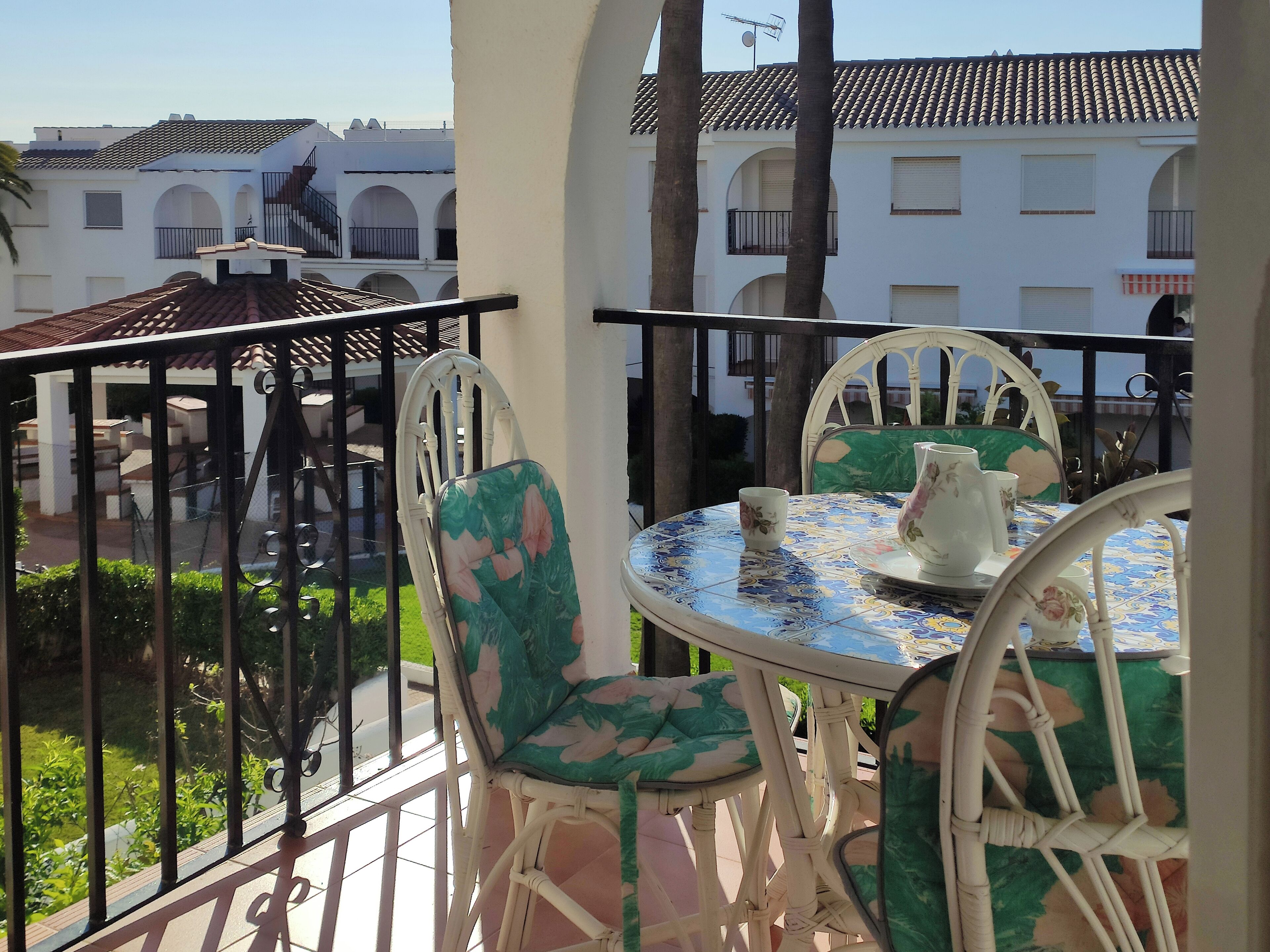 Un balcon avec des fauteuils en osier blanc et une table, donnant sur une cour intérieure bordée d’immeubles et de verdure.