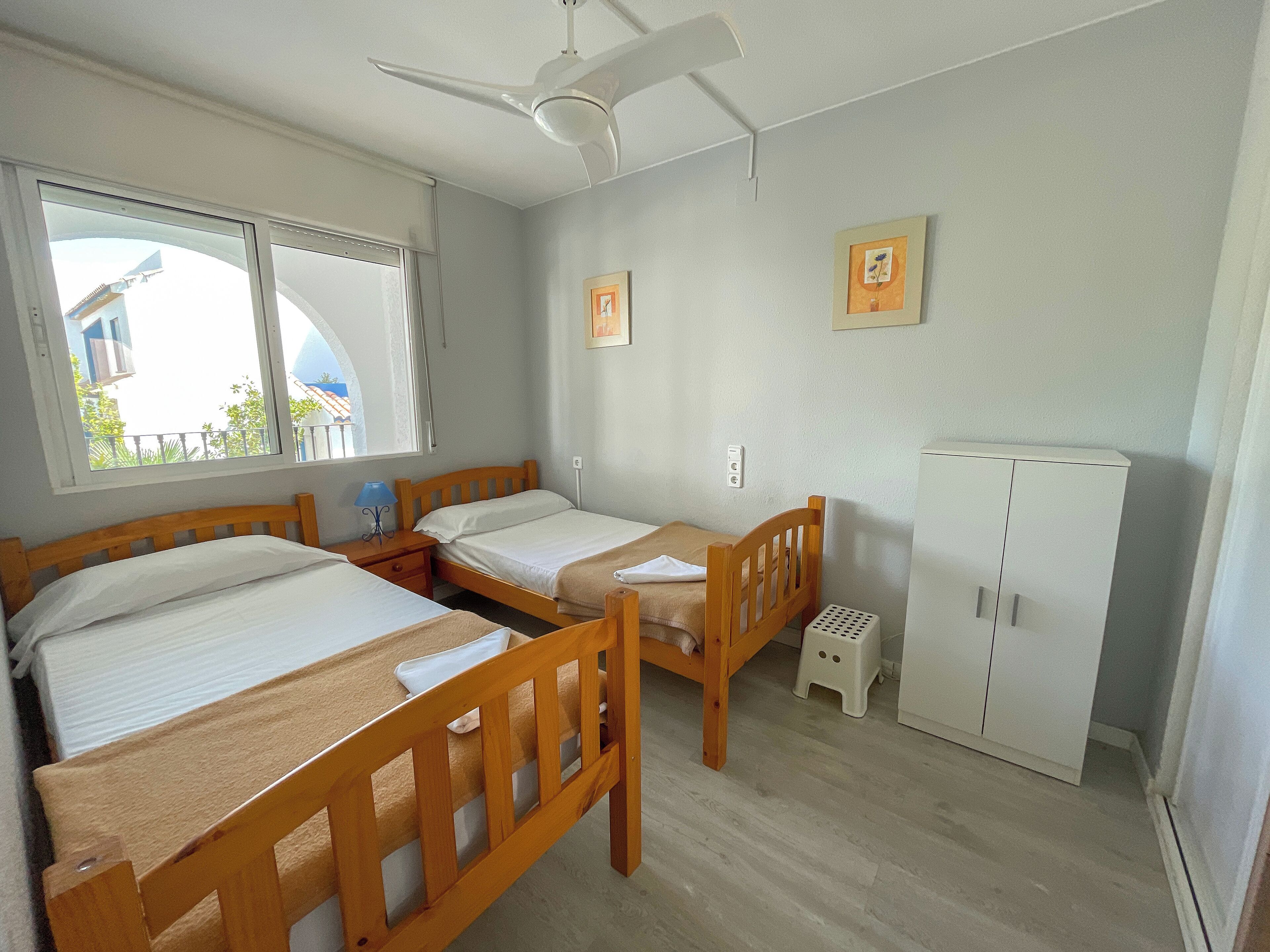 Ein Zimmer mit zwei Holzbett, einem Deckenventilator, einem weißen Kleiderschrank und einem Fenster mit Blick auf Gebäude.