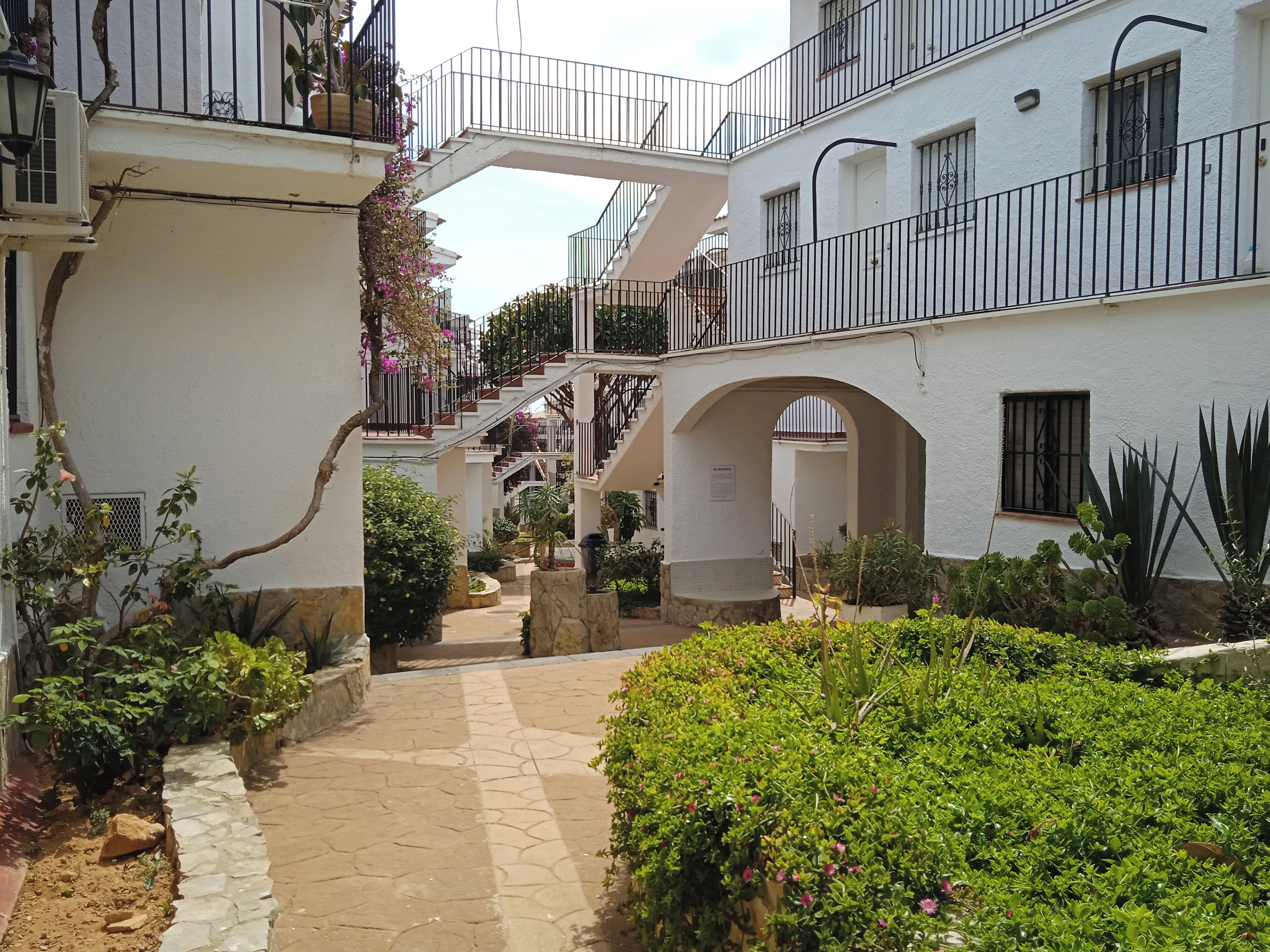 Un cortile con un vialetto in pietra, edifici bianchi con balconi e diverse piante.