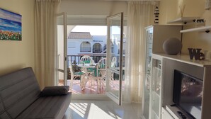 Uma sala de estar com sofá, TV e varanda com mesa e cadeiras.