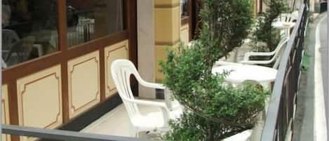 A balcony with potted plants and white plastic chairs, overlooking a street with buildings.