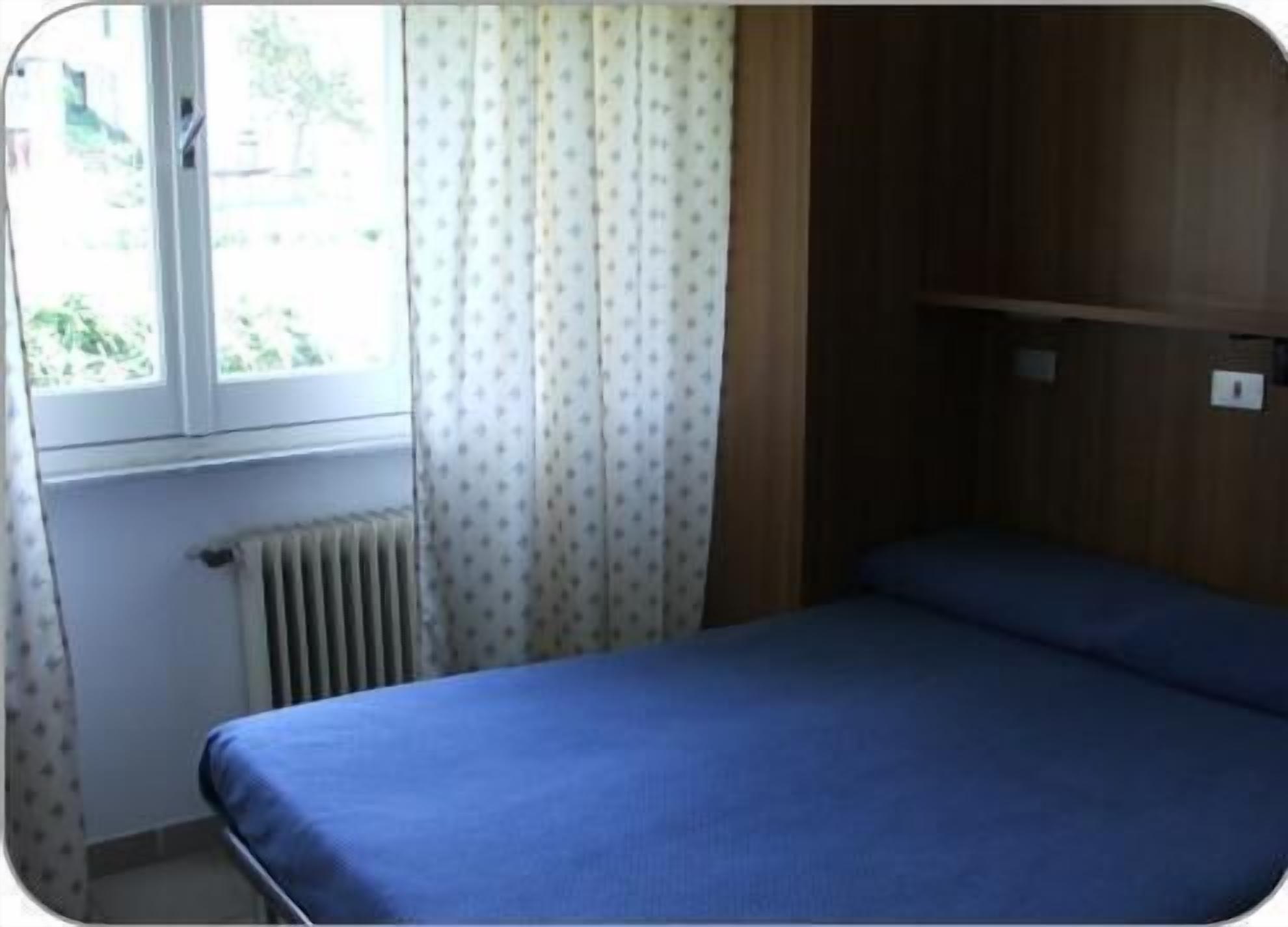 A small, single-bed room with a window, a radiator, and a wooden panel wall.