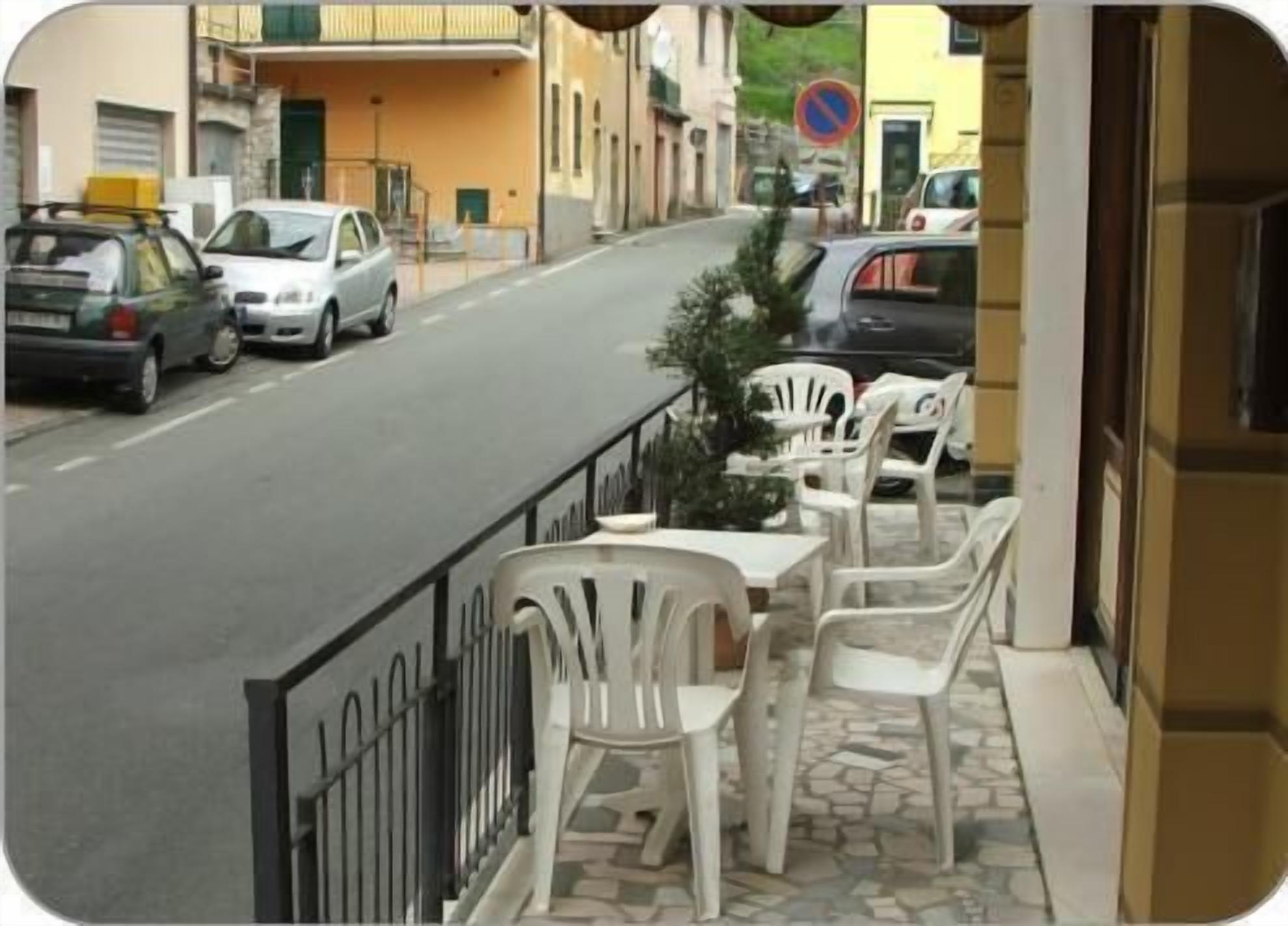 A street view with parked cars, a no parking sign, and outdoor seating with white plastic chairs and tables.